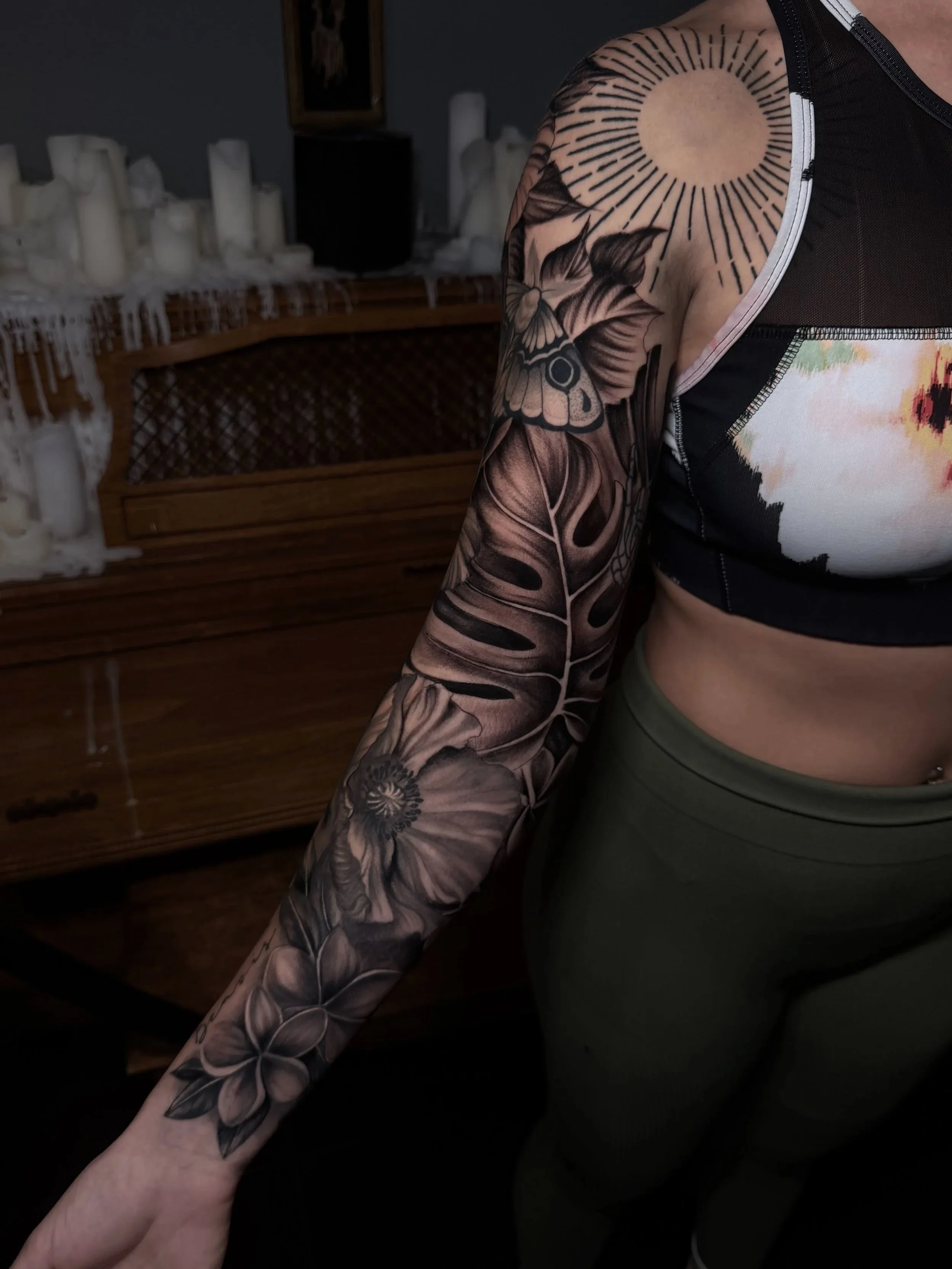 Close-up of a person’s floral and butterfly tattoo sleeve, wearing a black-and-white top in a dimly lit room.