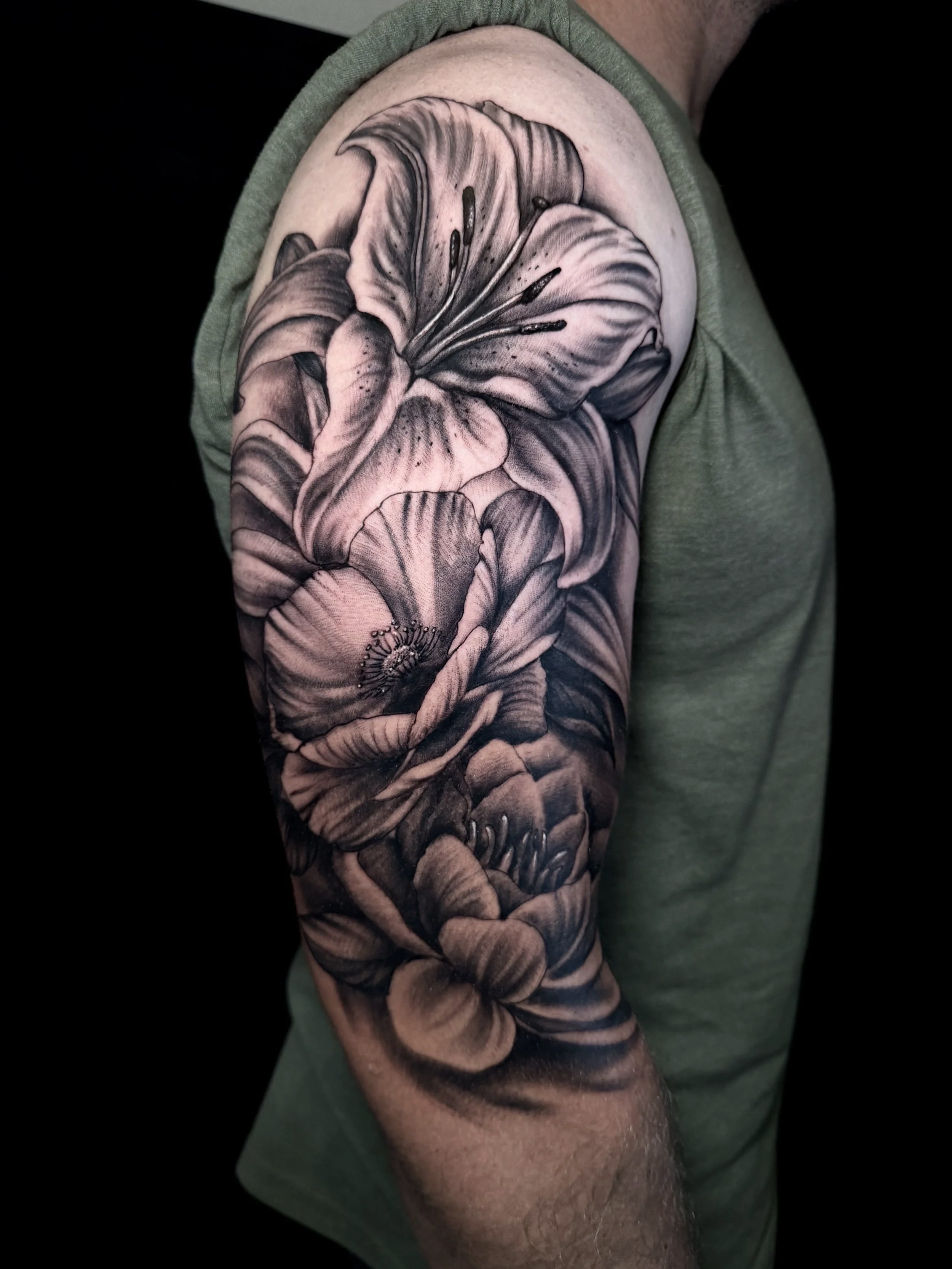 Black-and-gray botanical sleeve tattoo featuring detailed lilies and leaves on an upper arm