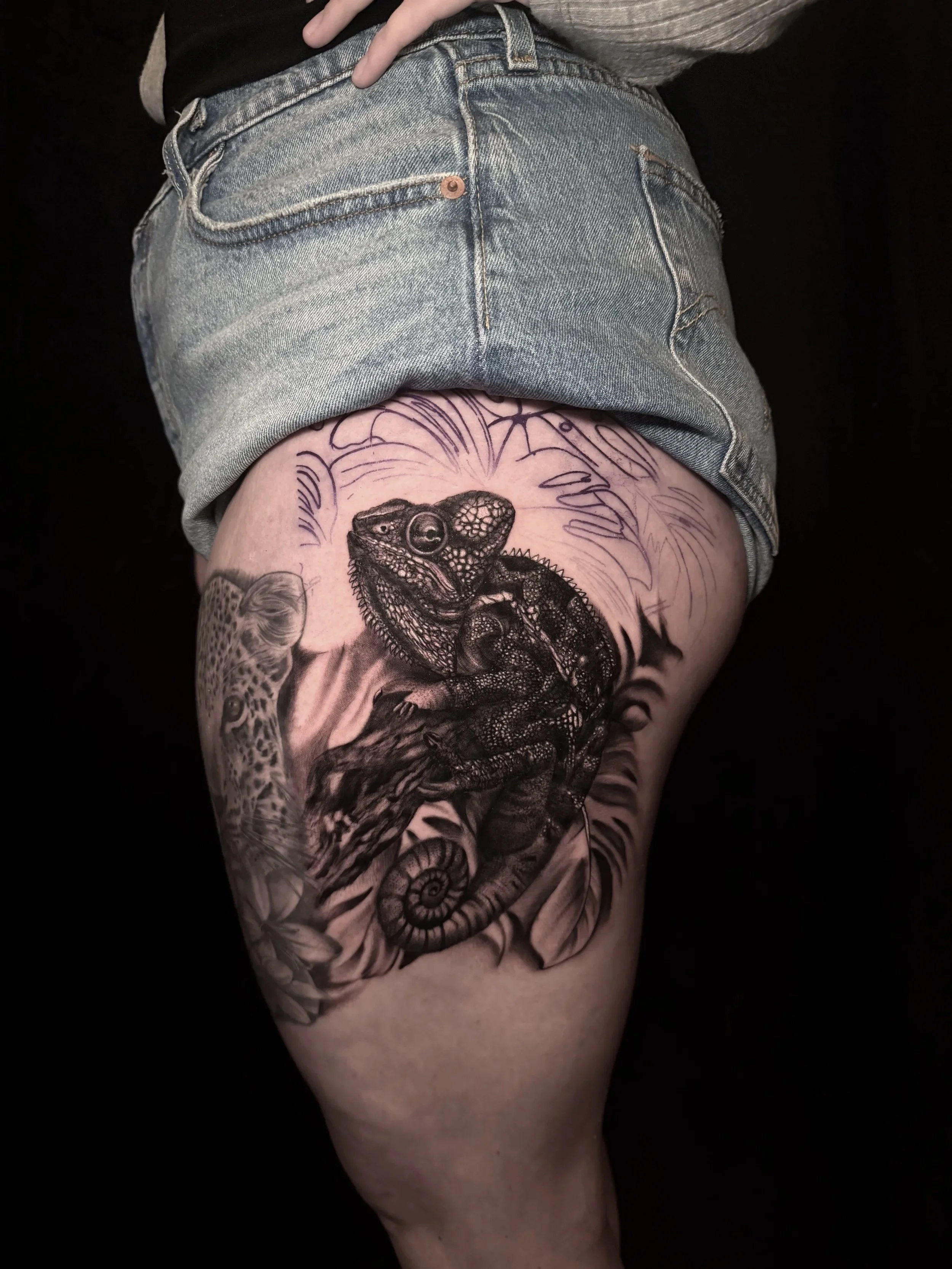 Close-up of an upper thigh with a detailed black-and-gray lizard tattoo surrounded by floral and botanical designs, partially covered by denim shorts.
