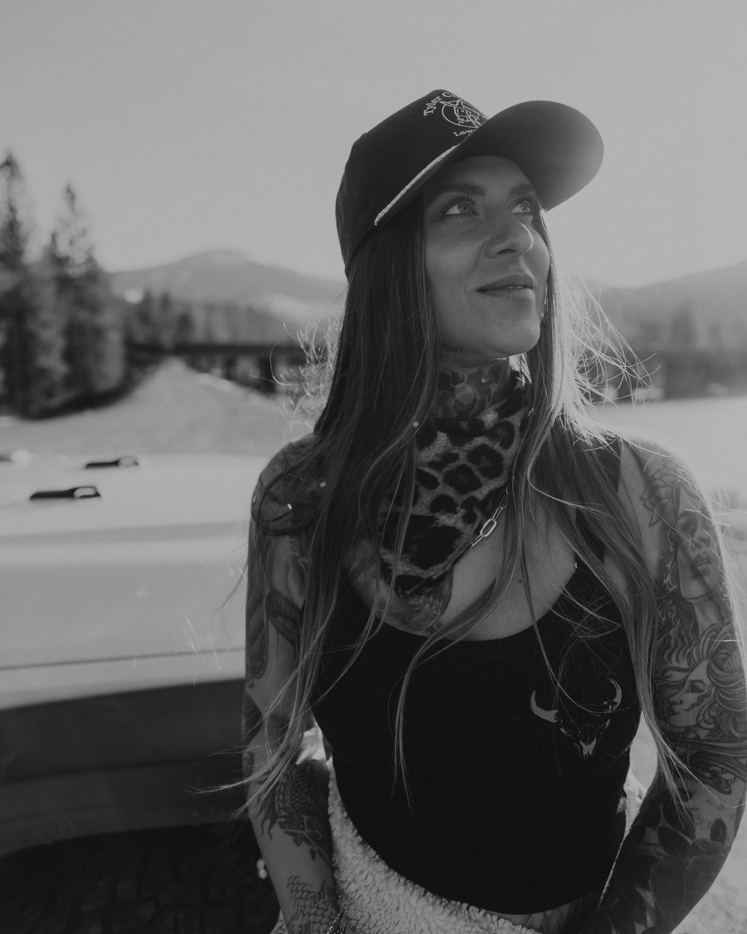 Black-and-white portrait of a tattooed woman wearing a baseball cap and scarf outdoors.