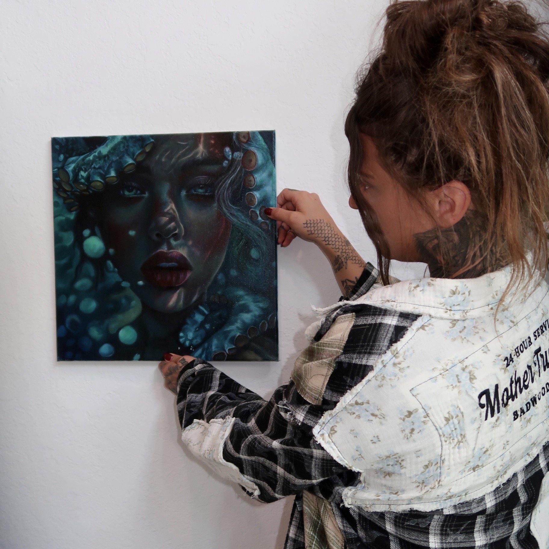 Tattooed woman in a plaid shirt examining a dark underwater-themed portrait painting on a wall