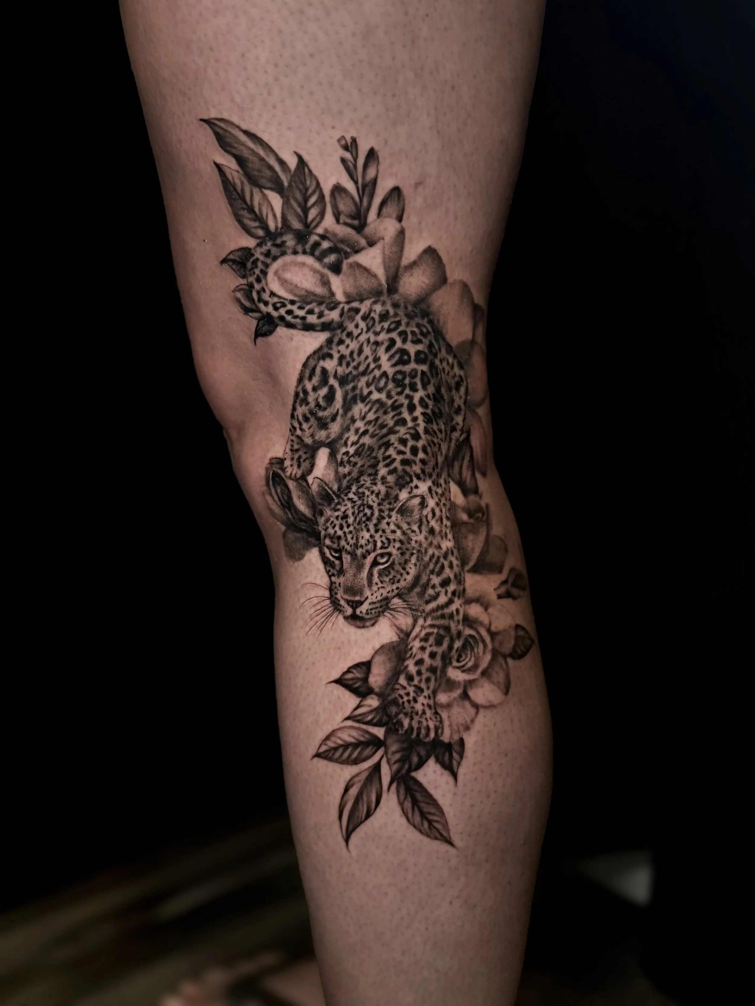 Detailed black-and-gray leopard tattoo surrounded by flowers and leaves on a leg