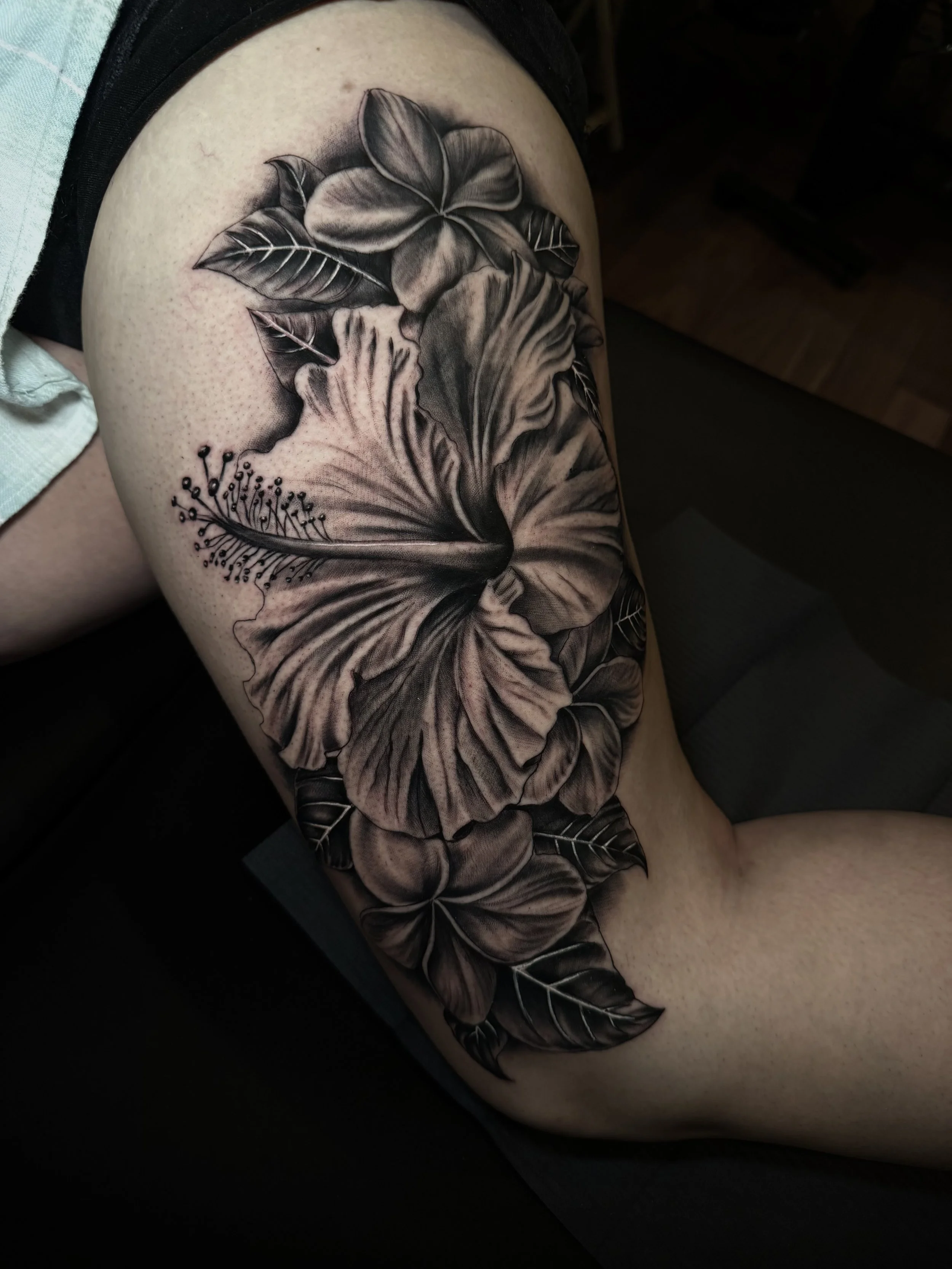 Black-and-gray floral tattoo featuring large blooming flowers, leaves, and delicate sprigs on a person's thigh