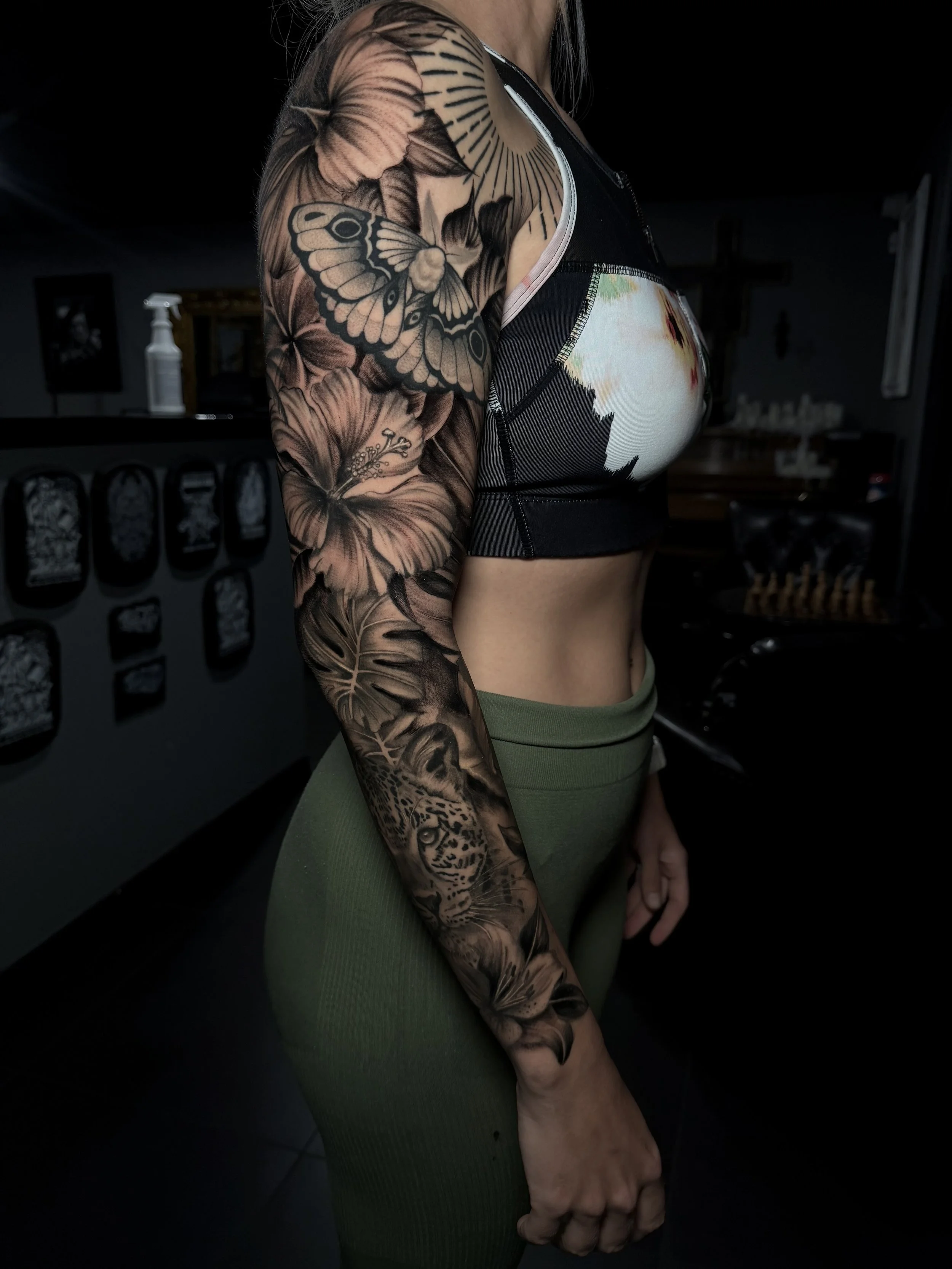 Person wearing a black crop top and green pants, showing a full-sleeve tattoo featuring flowers, a butterfly, leaves, and a leopard.