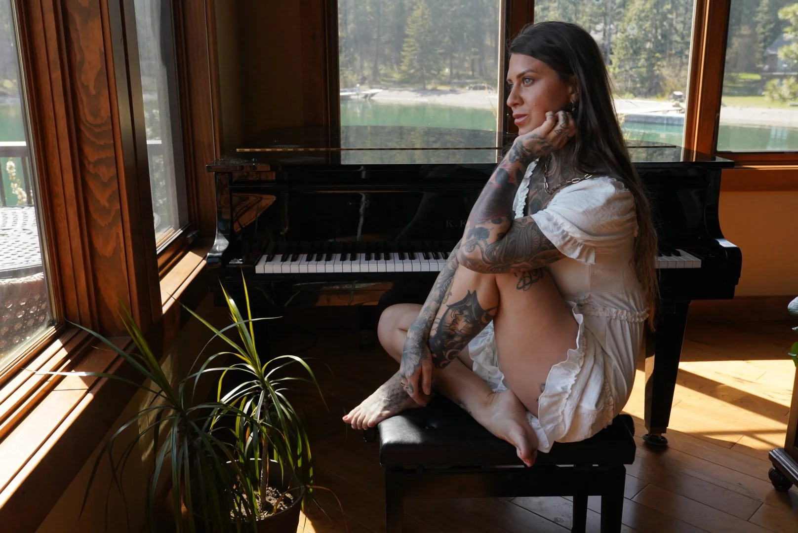 Tattooed woman in a white dress sitting beside a black grand piano in a sunlit room overlooking a lake