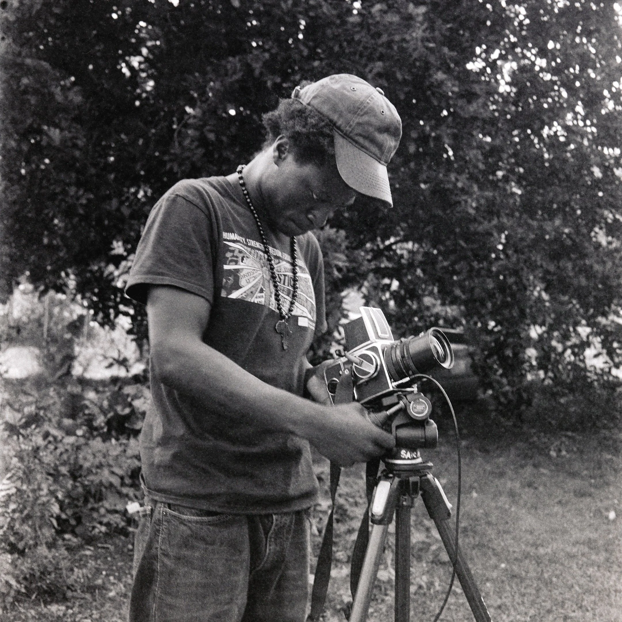 Black-and-white photo of a man operating a video camera on a tripod outdoors