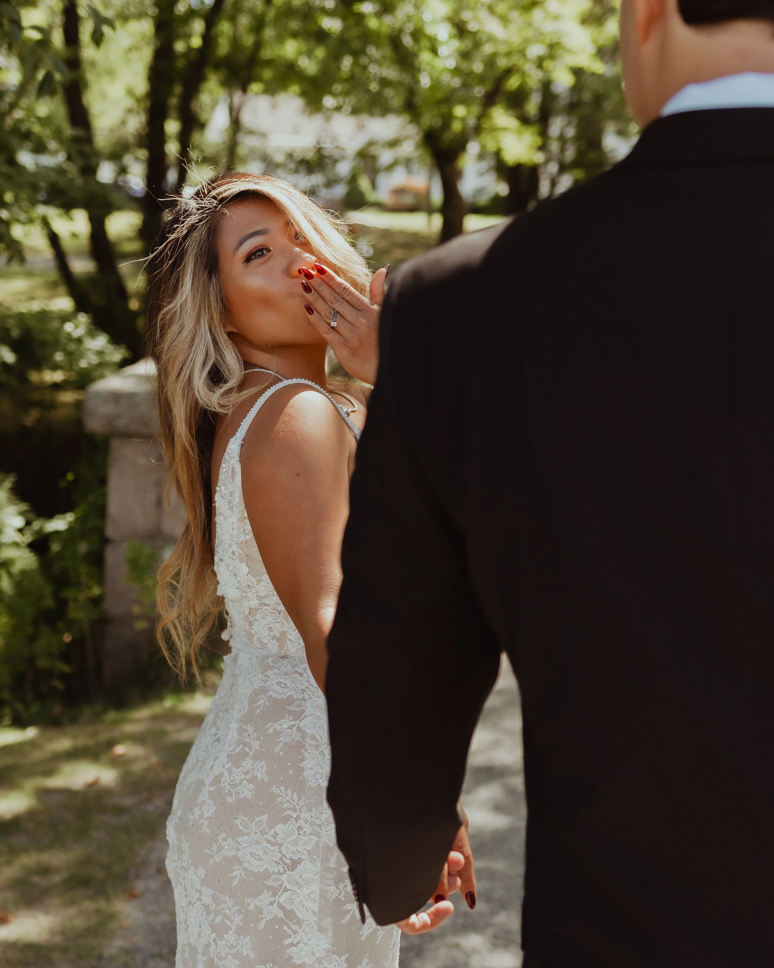 Bride in a lace wedding dress covers her mouth while walking hand in hand with her partner through a sunlit wooded park.