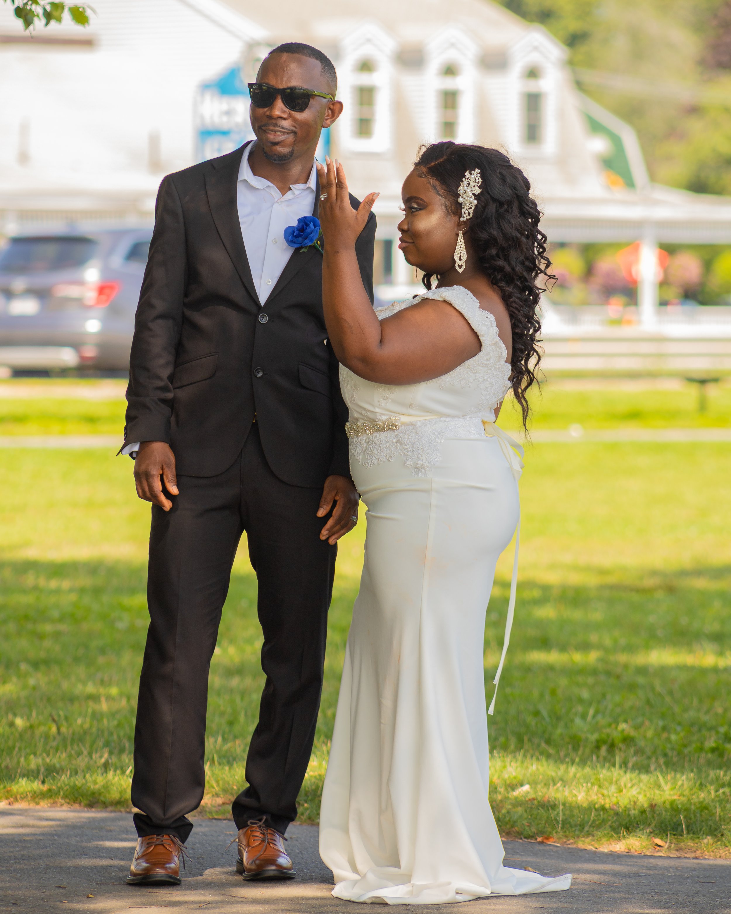 A image of Eric and Florence after their wedding ceremony. They are in a park, standing together, and Florence has her hand up to show off her ring and Eric stands happy by her side with sunglasses on and in a black suit with blue flowers. s
