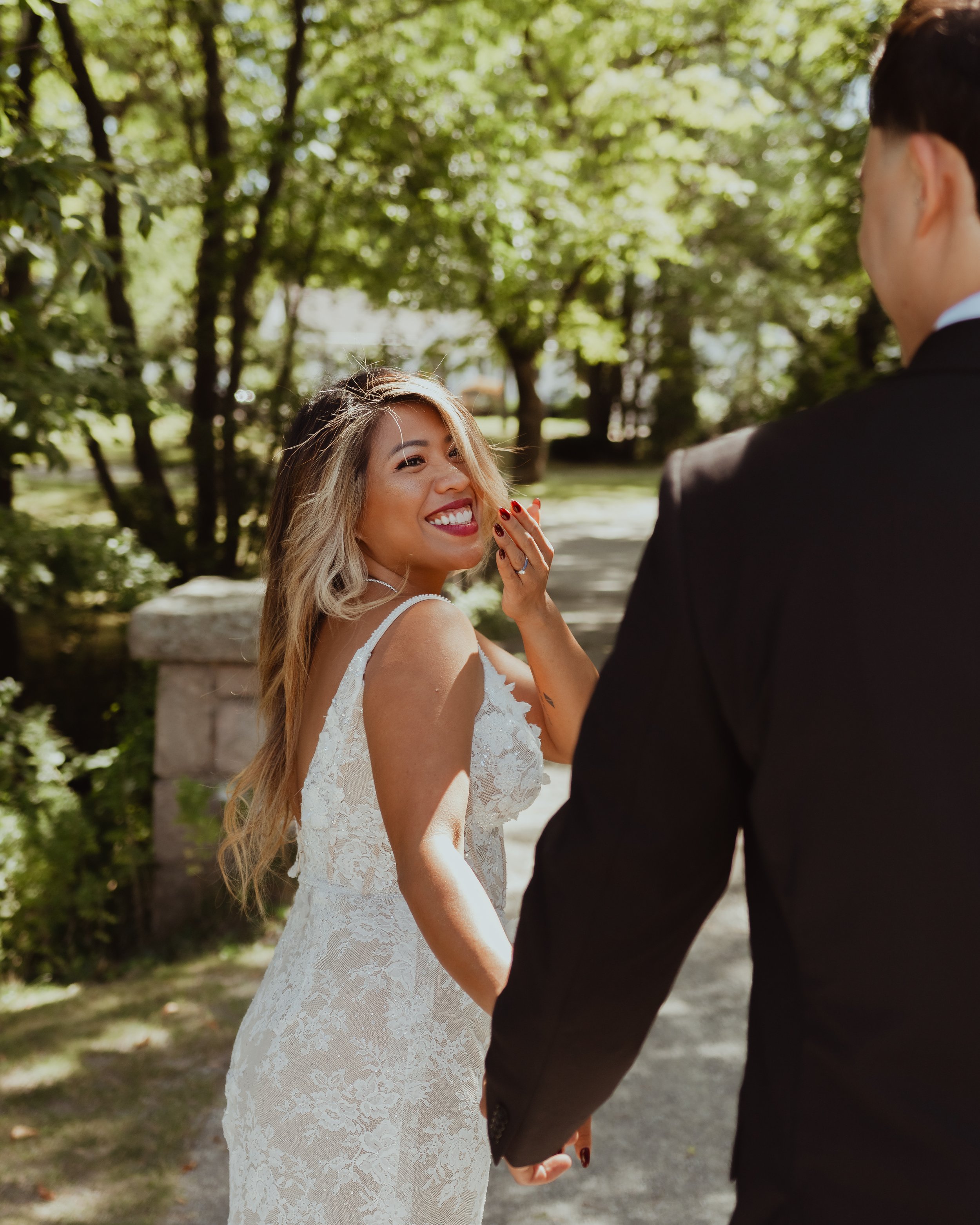 Smiling bride in a white lace dress holding hands with her groom on a sunlit wooded path