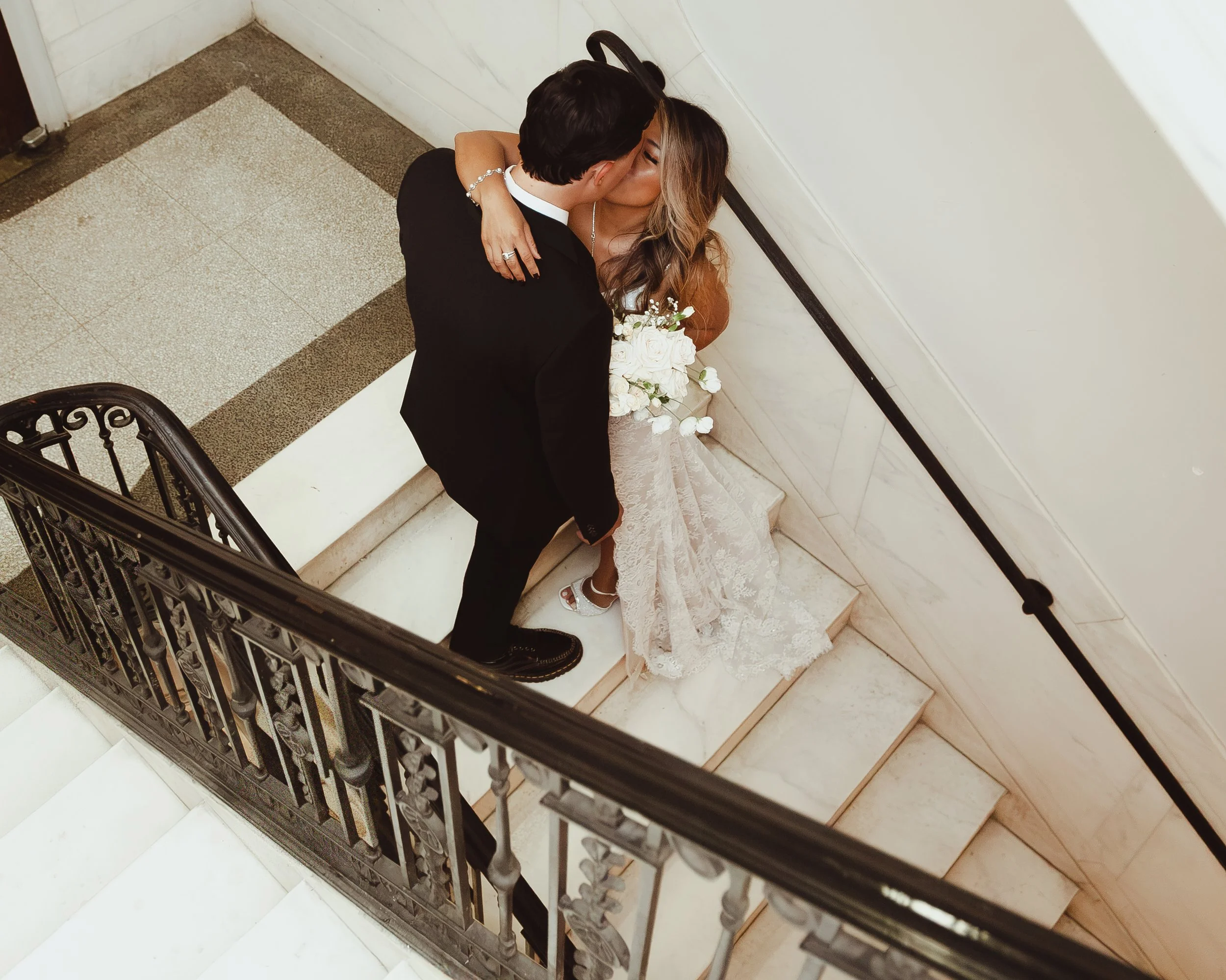 Bride and groom kissing on a marble staircase, with the bride holding a white wedding bouquet