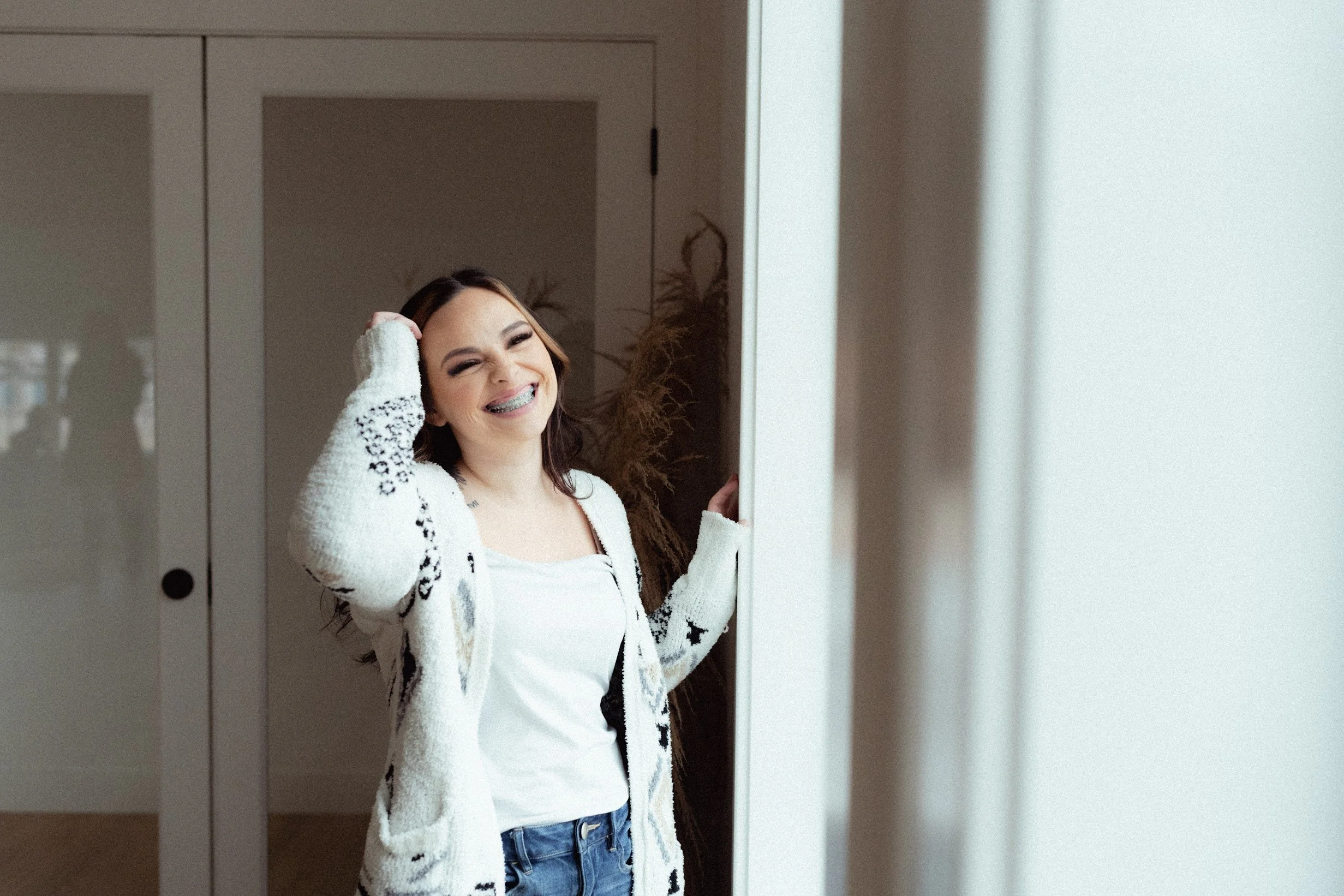Smiling woman with braces wearing a fuzzy white cardigan and jeans, standing in a doorway.