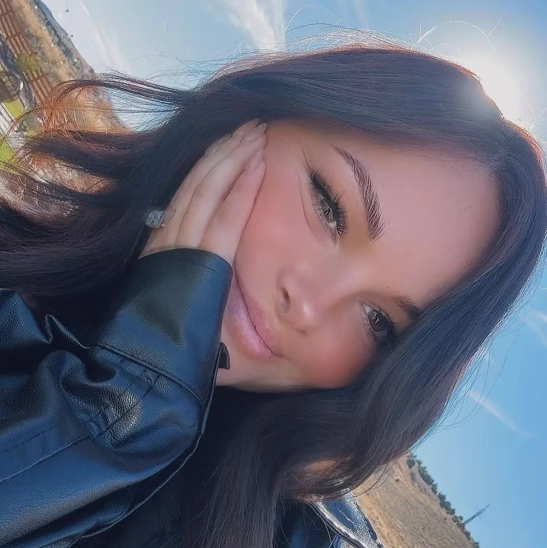Close-up outdoor selfie of a woman with long dark hair wearing a black leather jacket, hand resting on her cheek