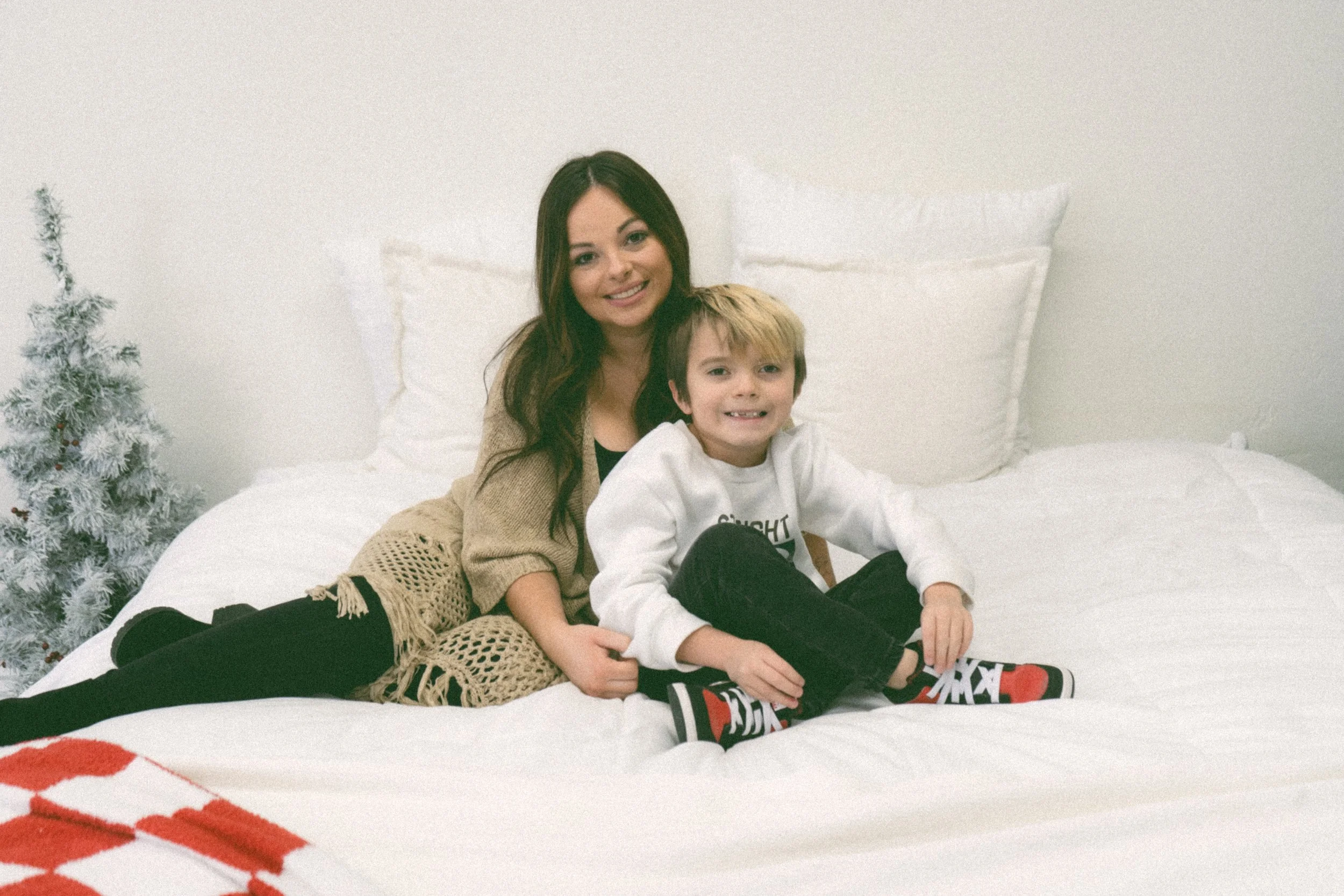 Smiling mother and young boy sitting on a white bed beside a small flocked Christmas tree.