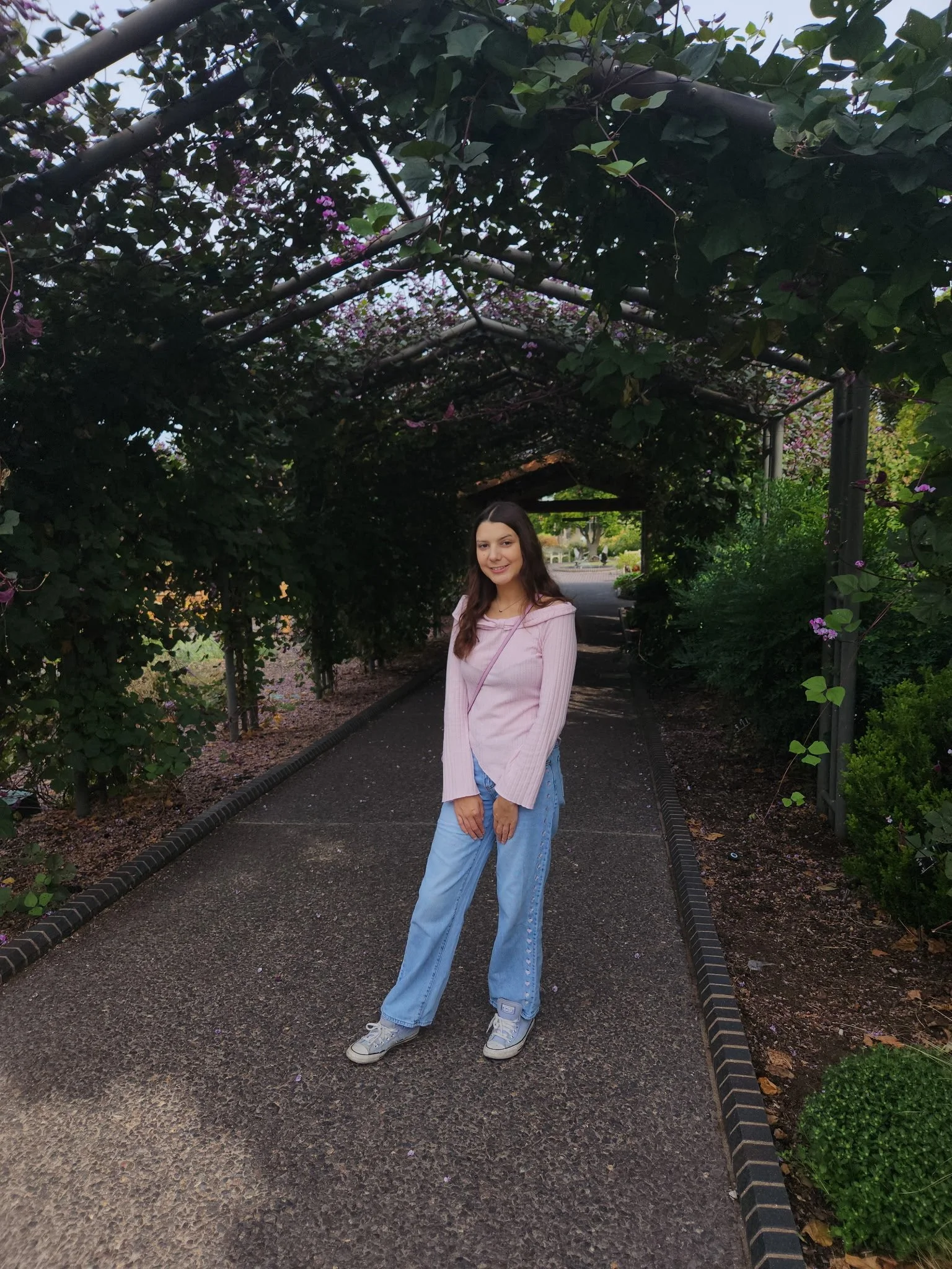 Woman in a pink top and jeans standing on a garden path beneath a flower-covered pergola