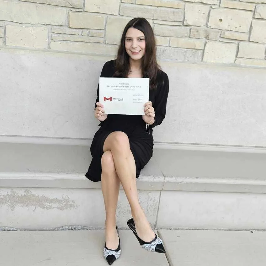 Smiling woman in a black dress holding an art award certificate while seated against a stone wall