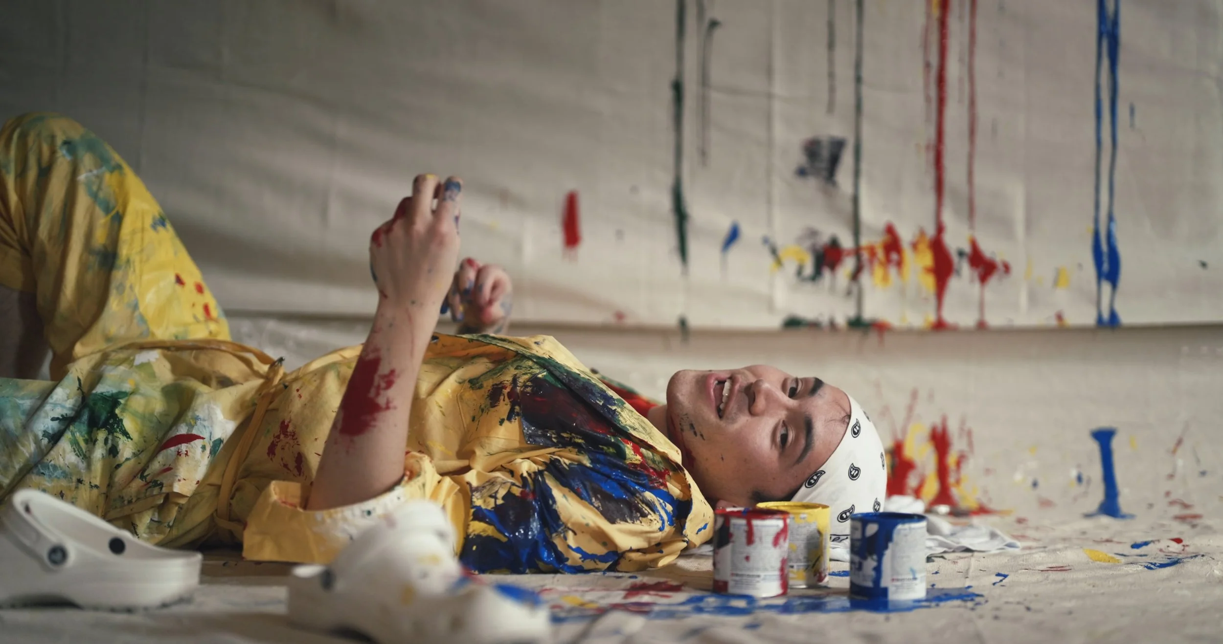 A painter covered in colorful paint lying on a studio floor, still from I Like Red!, a music video for Jean-Luc Swift