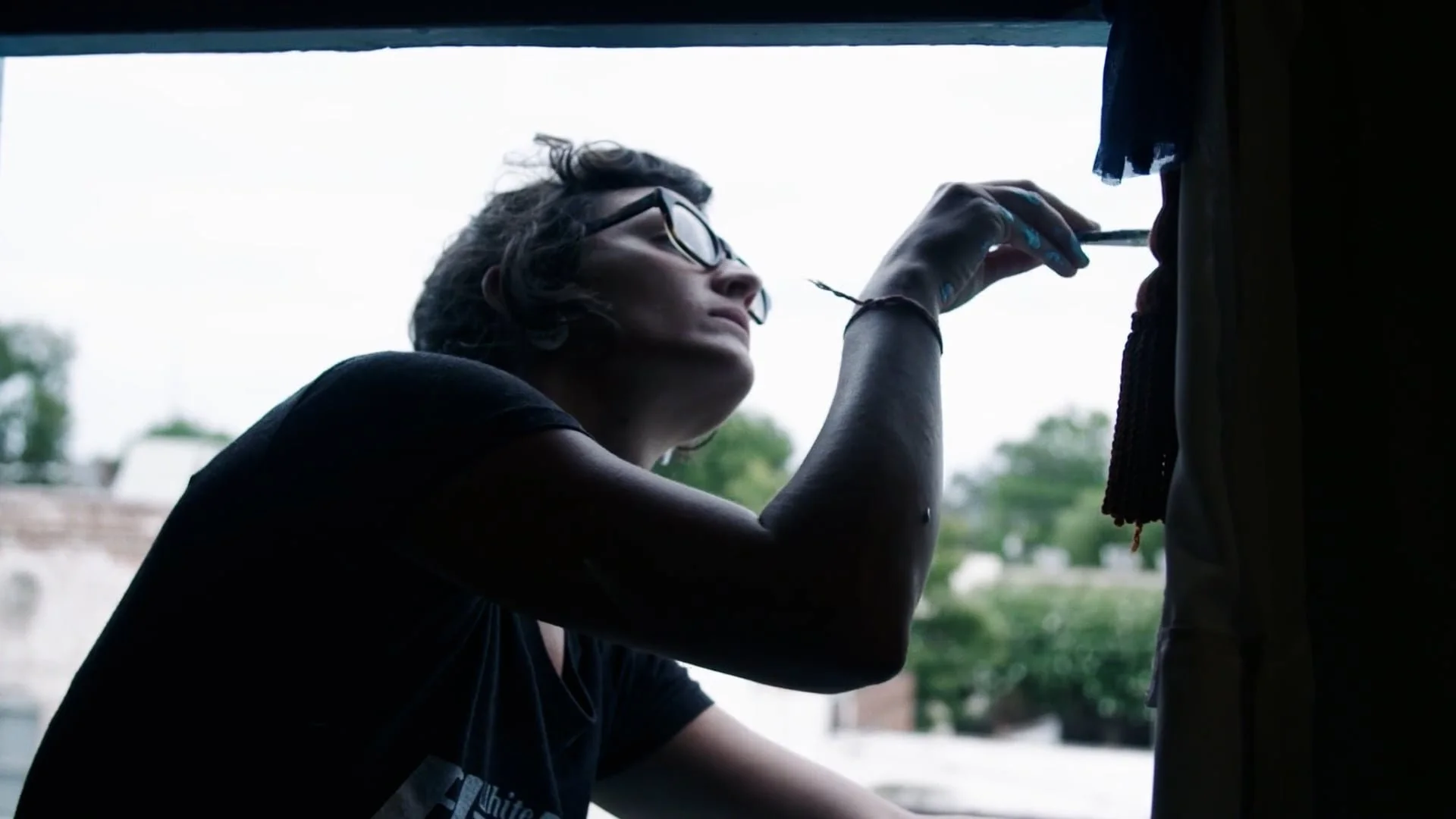 A woman in glasses adjusting curtains by an open window, still from Elsewhere, a film for the Elsewhere artist residency