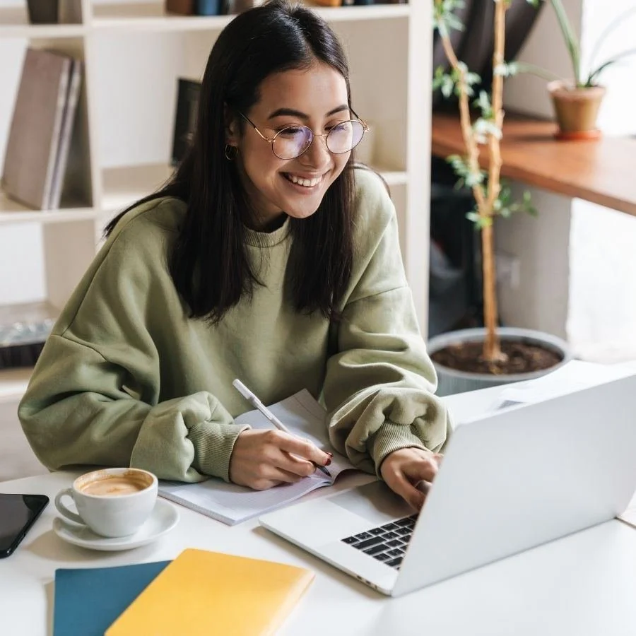 Smiling student taking notes beside a laptop at a bright desk