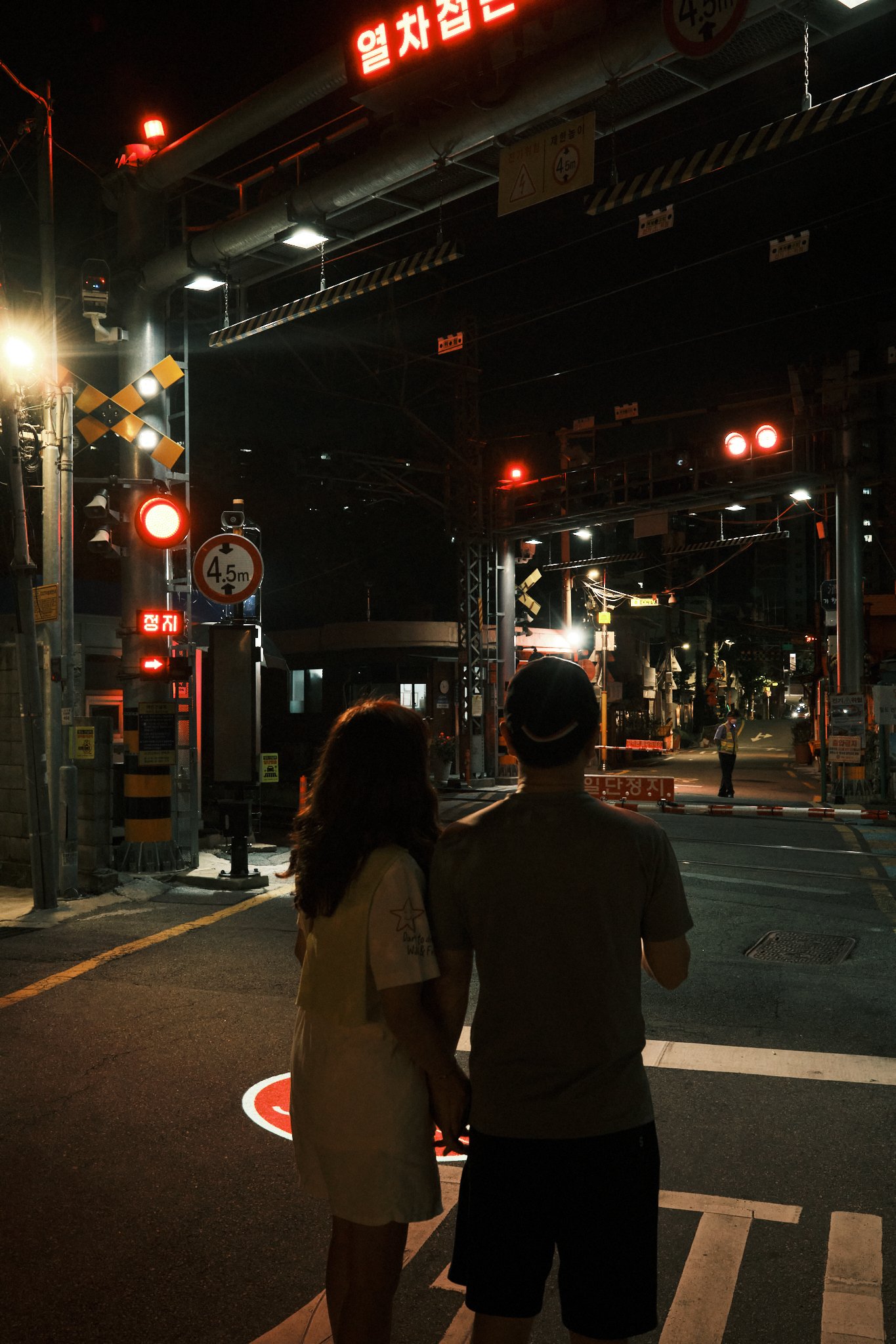 Two people wait at a nighttime Korean railroad crossing beneath illuminated Korean signs and red warning lights.