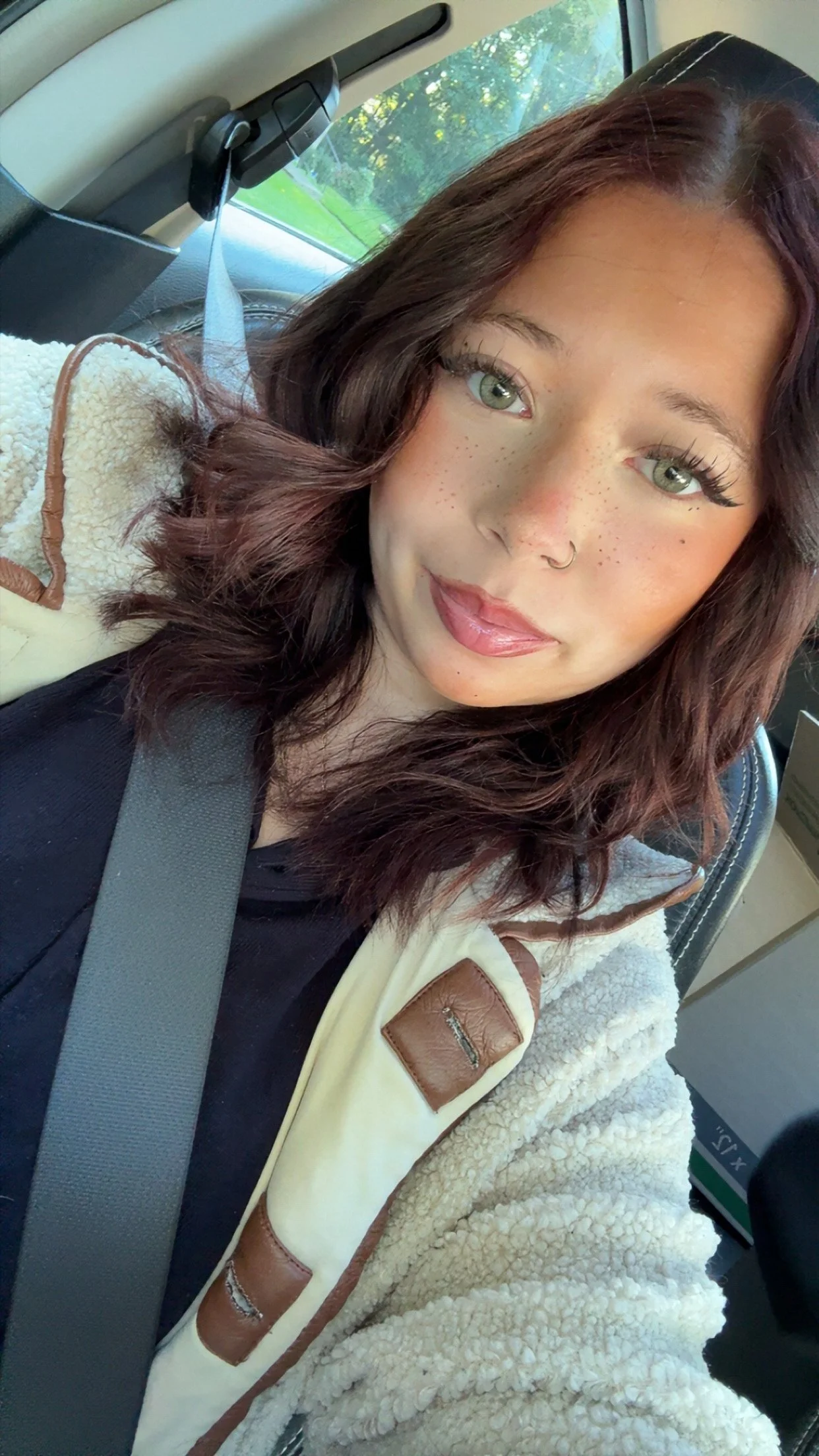 Selfie of a woman with long brown hair and green eyes wearing a sherpa-lined jacket and seat belt inside a car