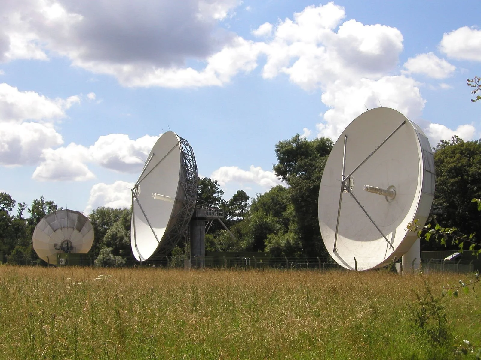Three large white radio telescope dishes in a grassy field beneath a partly cloudy blue sky