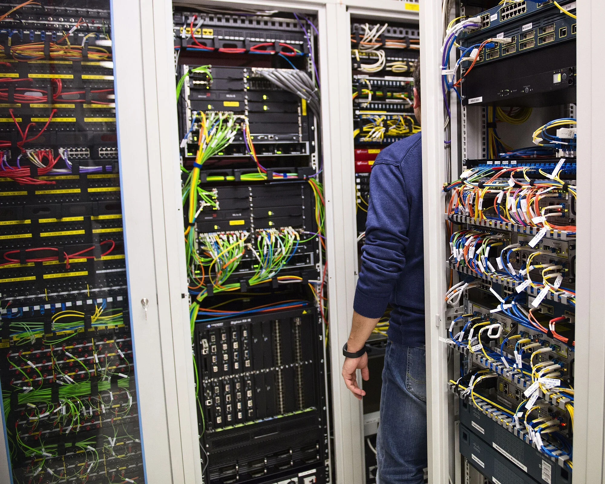 Technician standing among server racks filled with network switches, patch panels, and colorful cables in a data center
