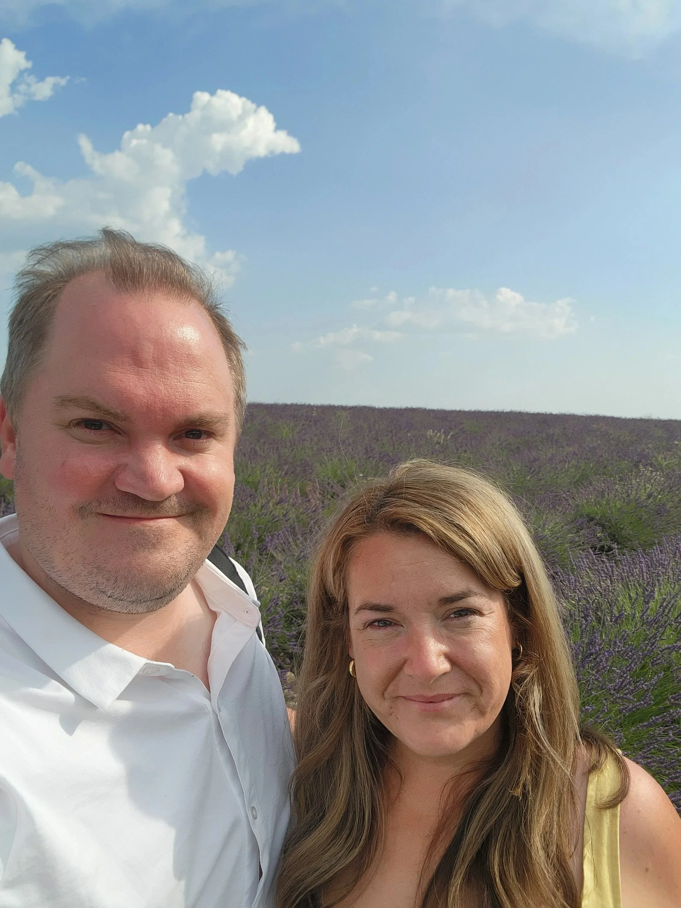 Smiling couple taking a selfie in a lavender field under a blue, cloud-filled sky