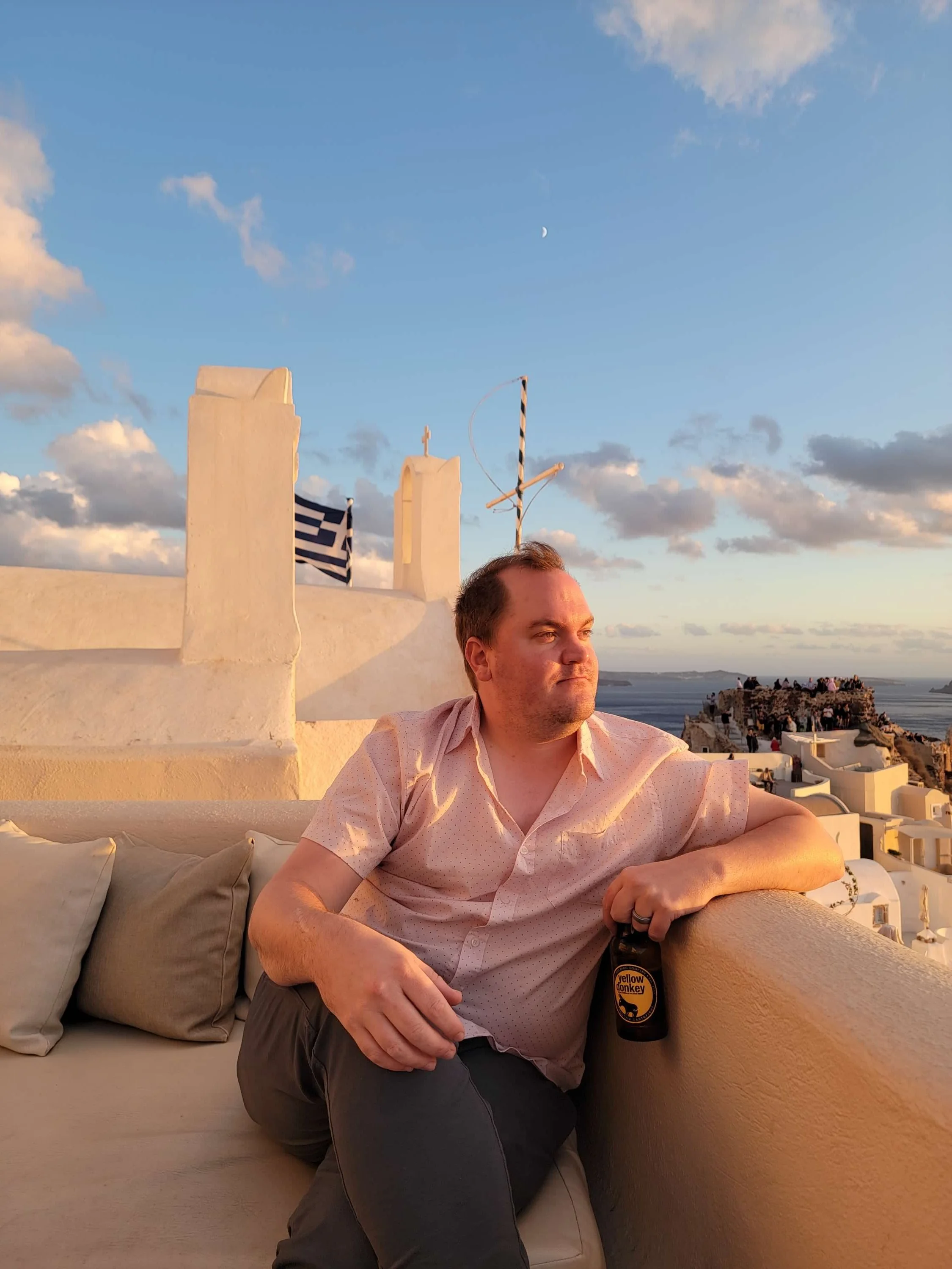 Man holding a beer bottle on a rooftop terrace overlooking the sea at sunset in Santorini, Greece