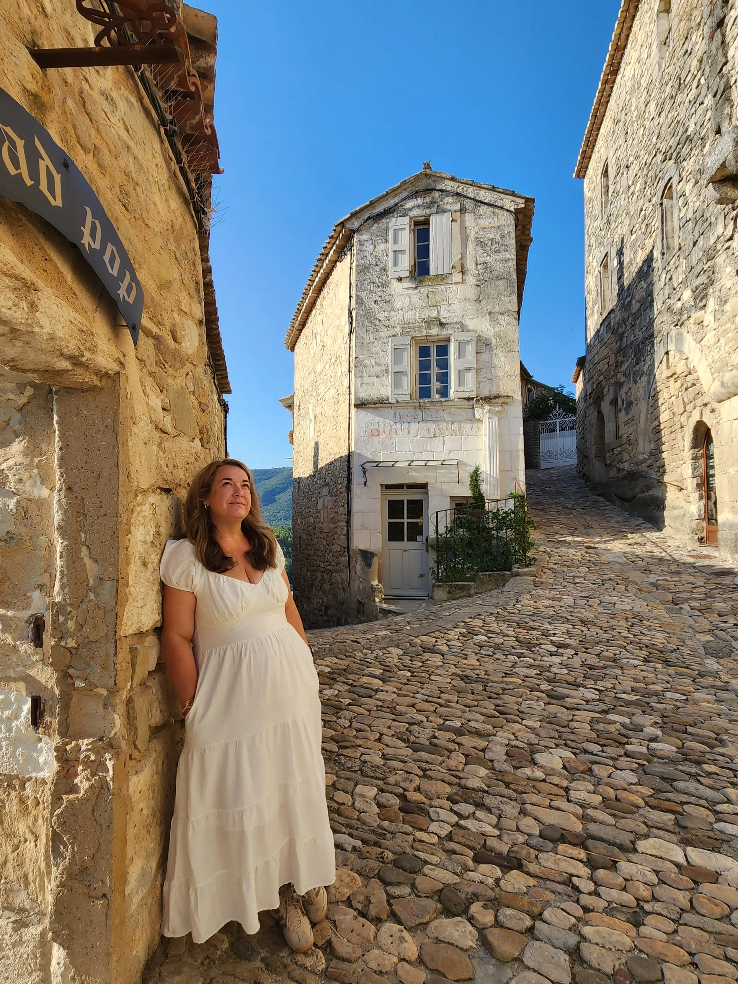 Woman in a white dress standing on a cobblestone lane between historic stone buildings under a clear blue sky