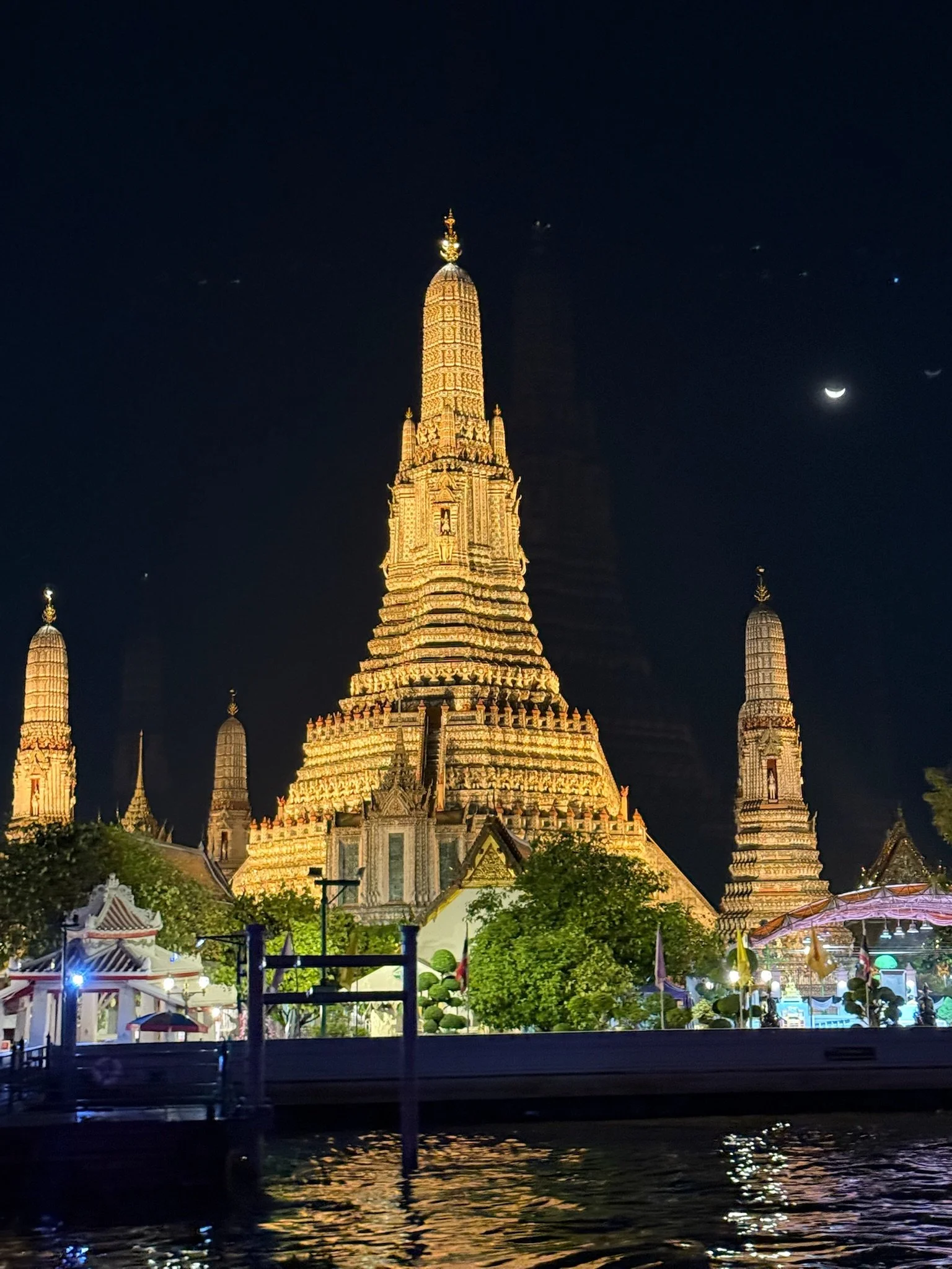 Wat Arun temple illuminated at night beside the Chao Phraya River in Bangkok, Thailand, with a crescent moon overhead