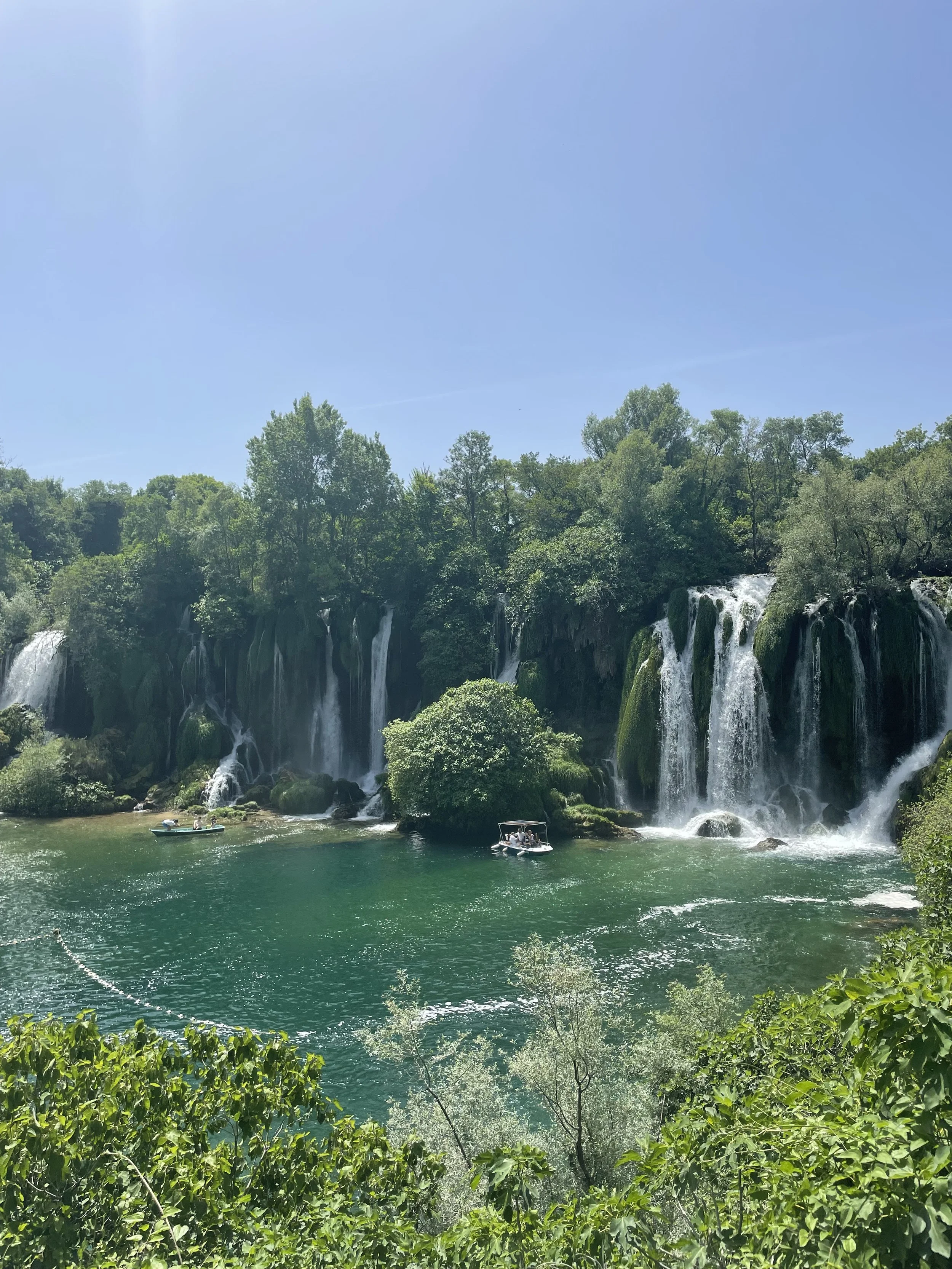 Scenic view of cascading waterfalls surrounded by lush green trees, with small boats on a turquoise pool.