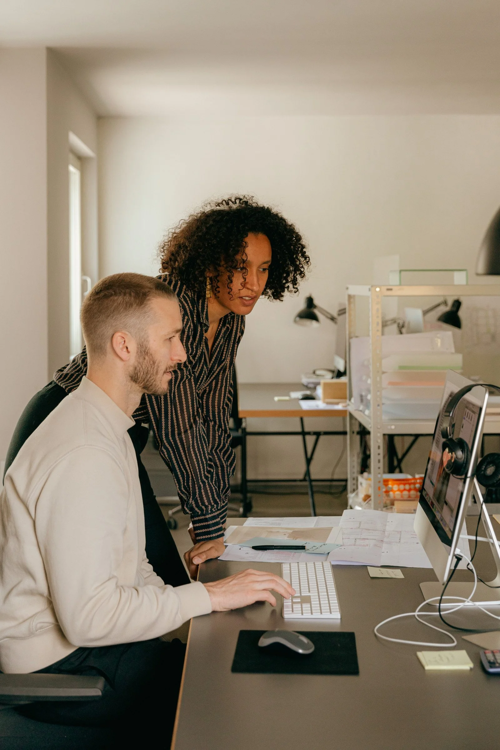man sits at computer and talks to a woman standing next to him