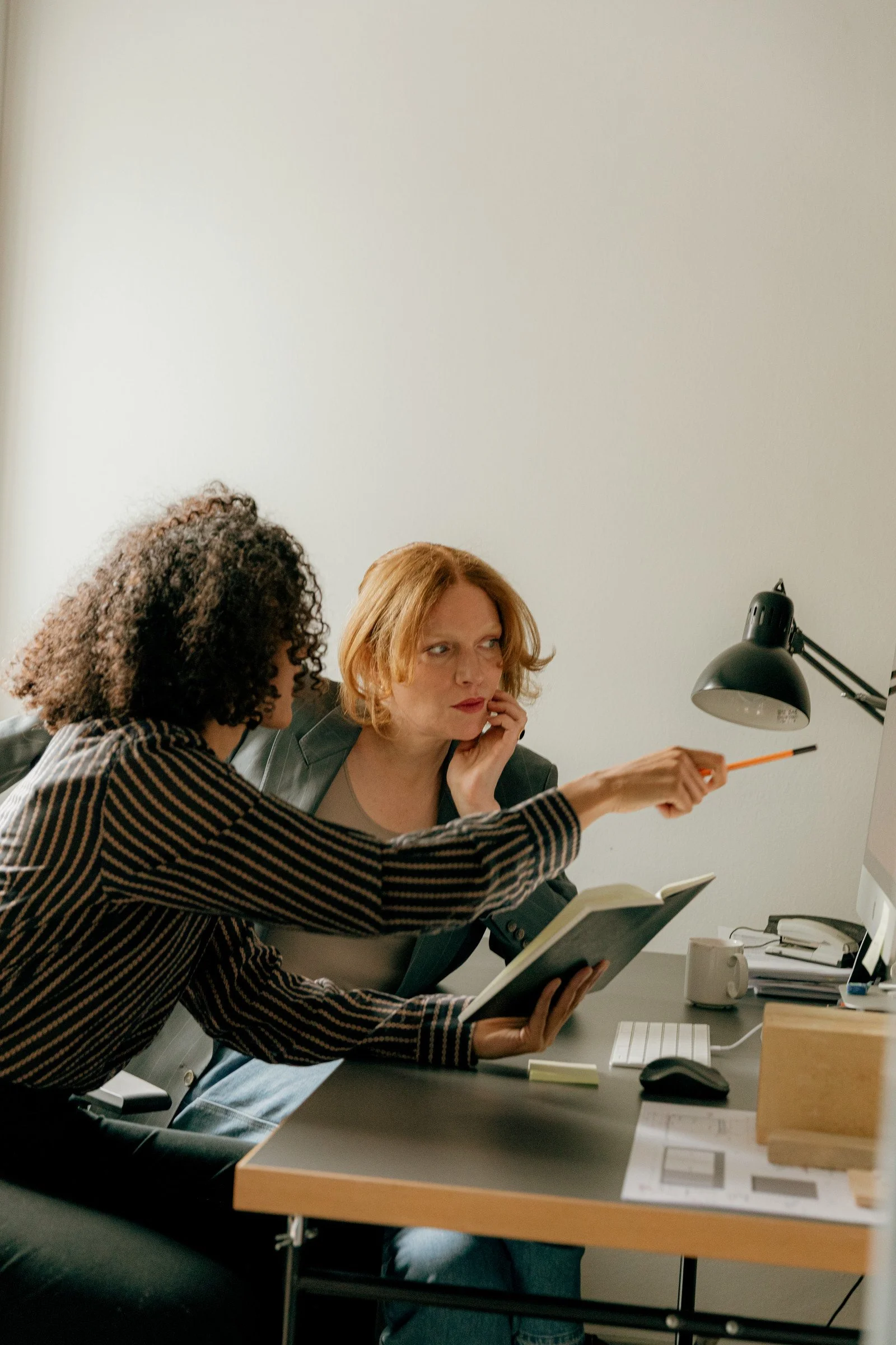 two women sit in an office space and discuss work by a computer