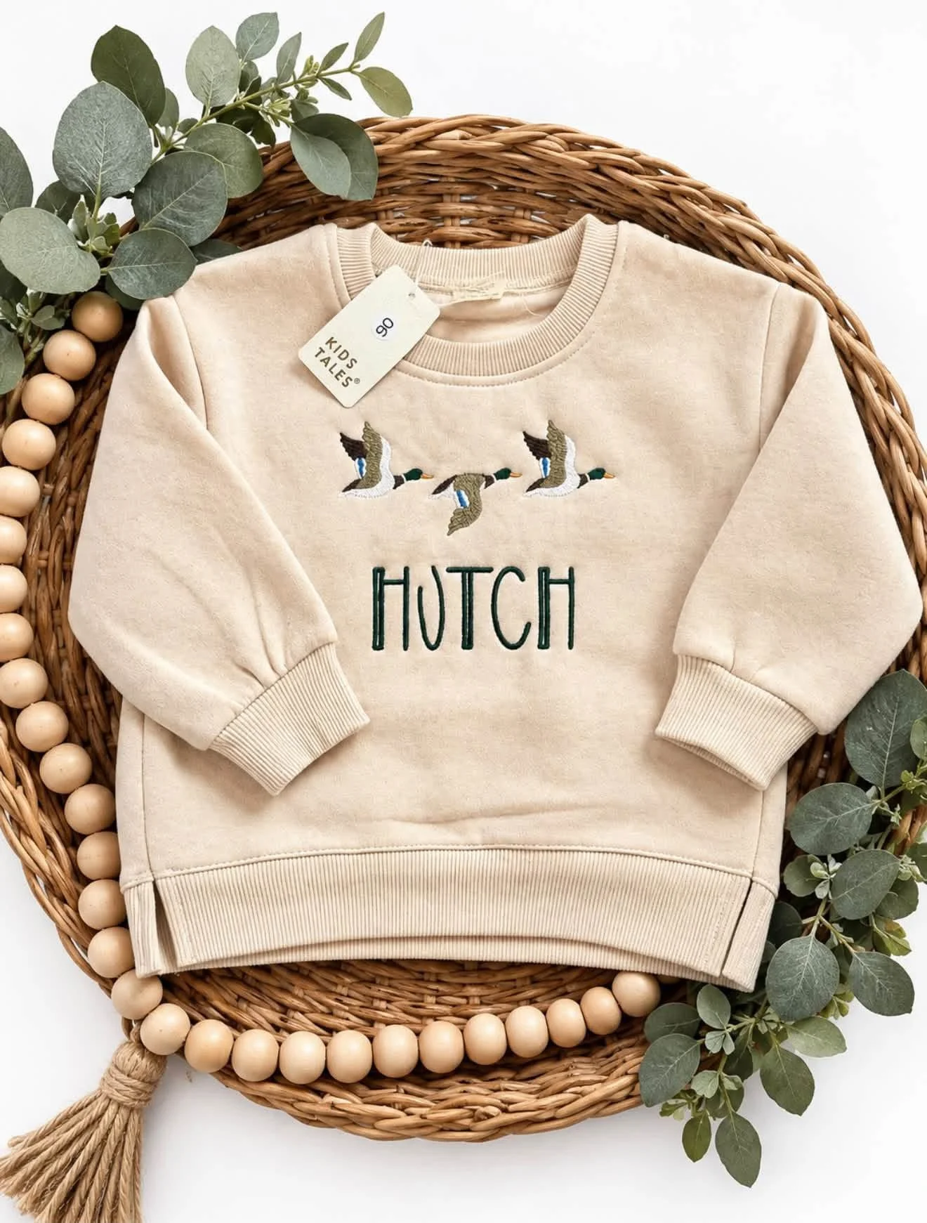 Cream kids' sweatshirt embroidered with flying geese and the word “HUTCH,” styled on a wicker tray with eucalyptus leaves.