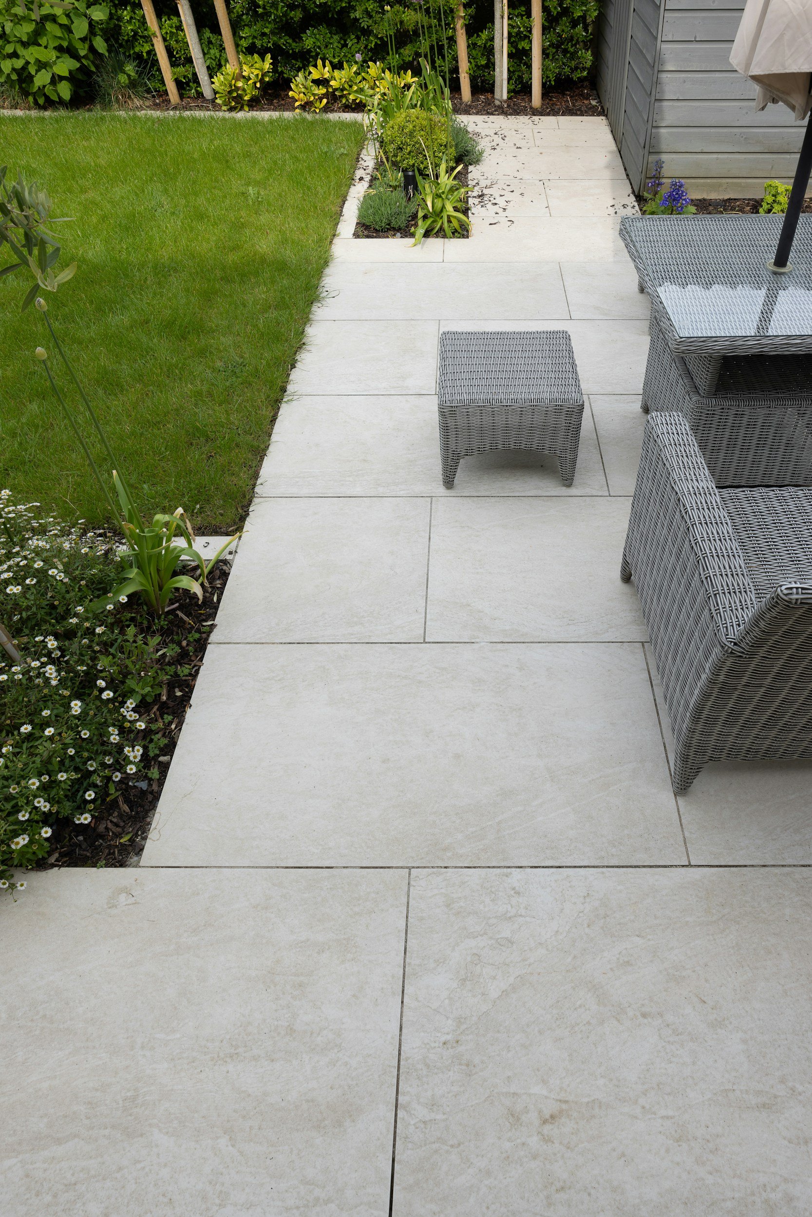 Gepflasterte Gartenterrasse mit grauen Rattan-Gartenmöbeln, Rasenfläche und bepflanzten Beeten neben einem Gartenhaus