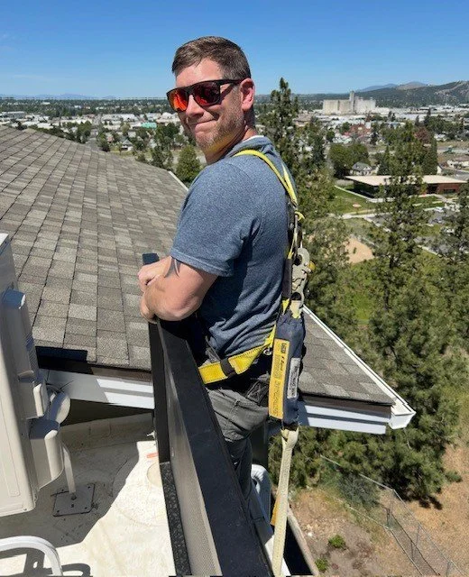 Roofer wearing safety harness and sunglasses stands on a rooftop overlooking a suburban neighborhood and mountains