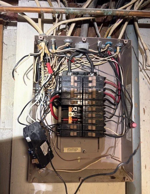 Open electrical breaker panel with exposed wiring, circuit breakers, and a loose black component