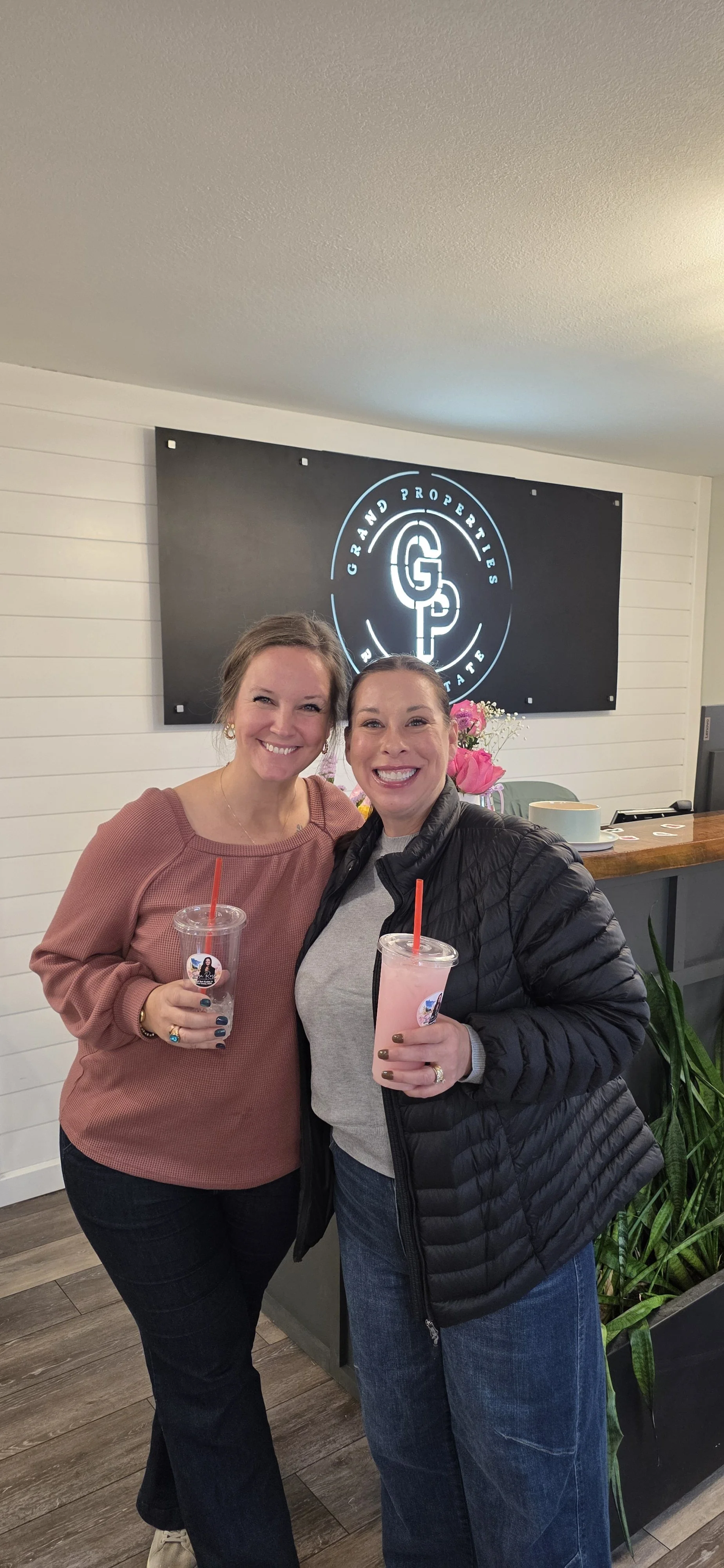 Two smiling women holding pink drinks in a café beneath a Great Properties sign, with flowers and a potted plant nearby.