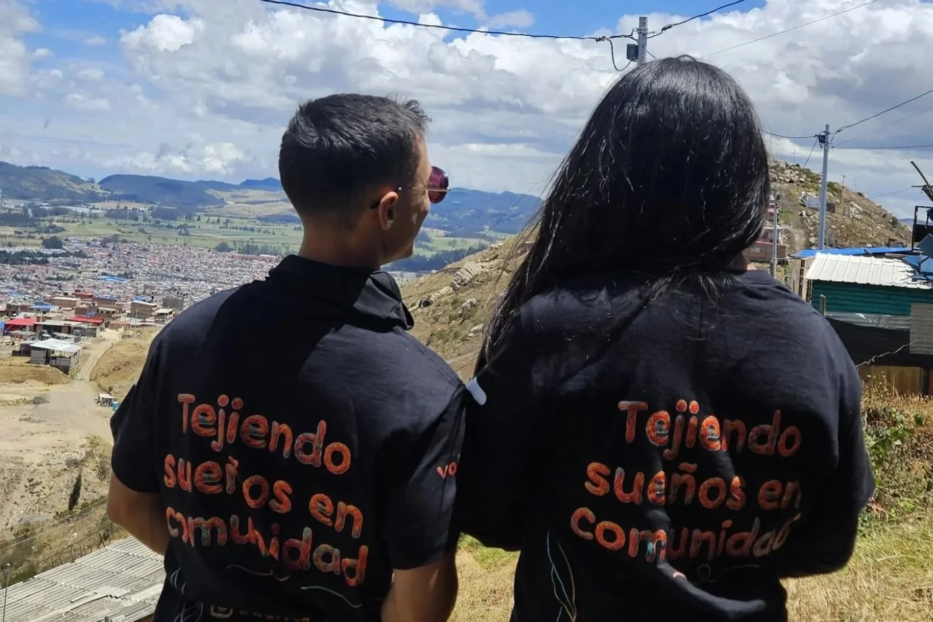 Dos personas con camisetas negras observan una ciudad andina rodeada de montañas y campos verdes.