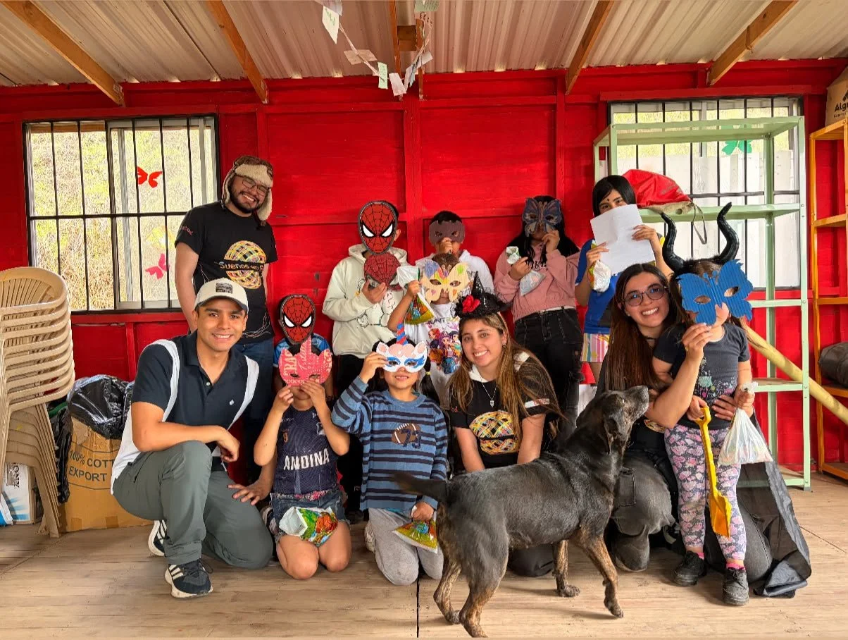 Grupo de personas con máscaras coloridas posa dentro de una cabaña roja junto a un burro.