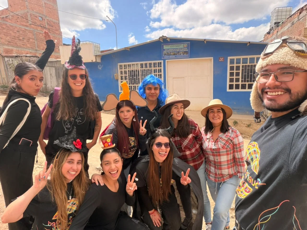 Grupo de amigos sonrientes disfrazados, tomándose una selfie frente a una casa azul en un día soleado.