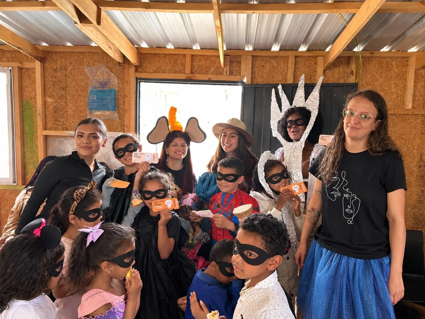 Grupo de niños y adultos disfrazados, con antifaces y accesorios de superhéroes, reunidos dentro de una cabaña.