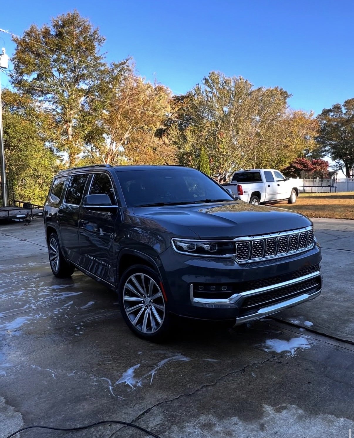 Dark blue Jeep Grand Cherokee SUV parked on a wet driveway with a white pickup truck and autumn trees in the background