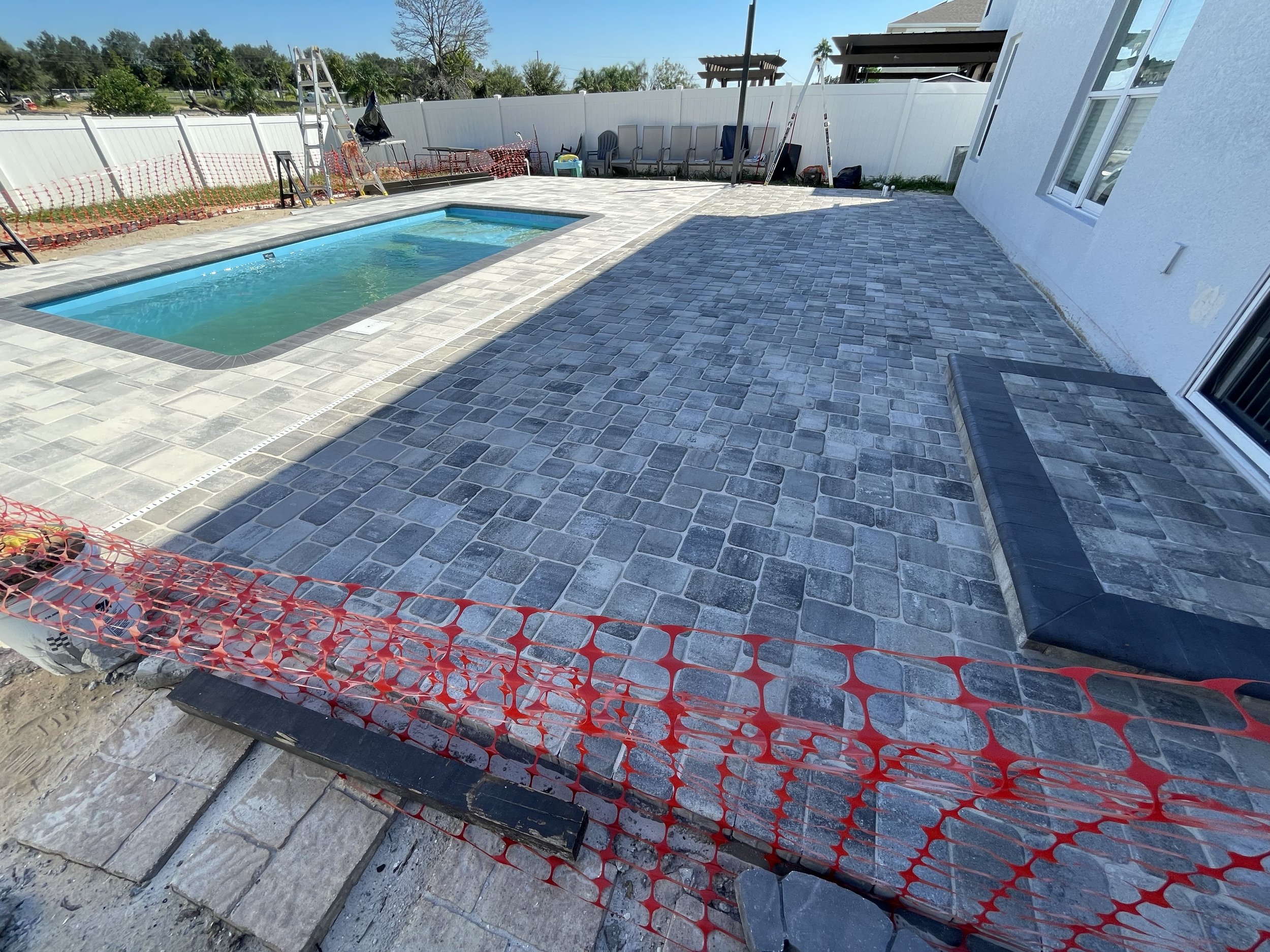 Backyard swimming pool surrounded by gray paver decking, white fencing, patio furniture, and construction safety fencing.