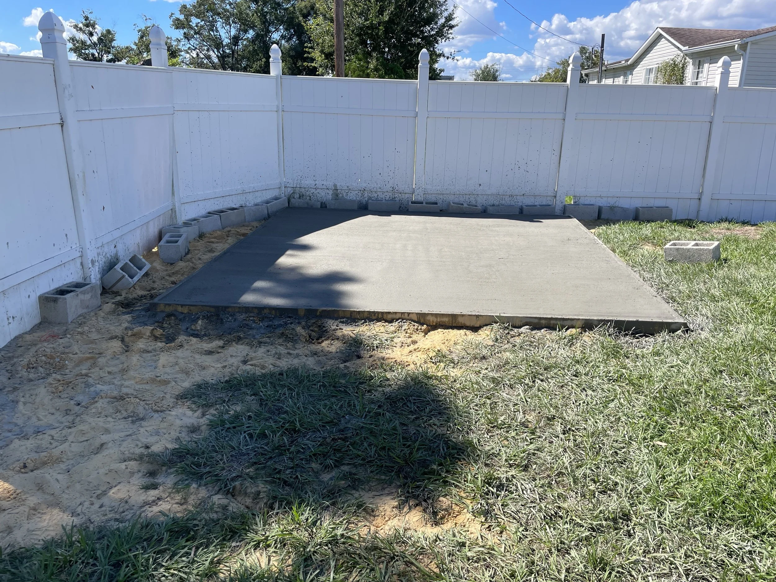 Newly poured concrete driveway or patio beside a white privacy fence, with cinder blocks, grass, and a house under a sunny blue sky