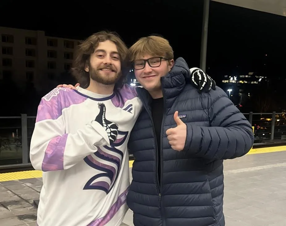 Two smiling young men pose together on a nighttime train platform; one wears a Seattle Kraken hockey jersey, and the other gives a thumbs-up in a navy puffer jacket.