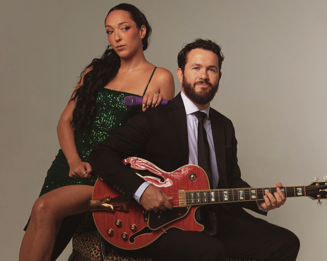 Woman in a green sequined dress poses behind a bearded man in a suit playing a red electric guitar.