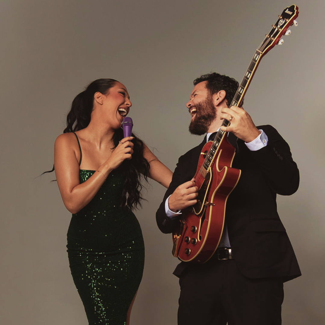 Singer in a sparkling green dress laughing with a suited guitarist playing a red electric guitar