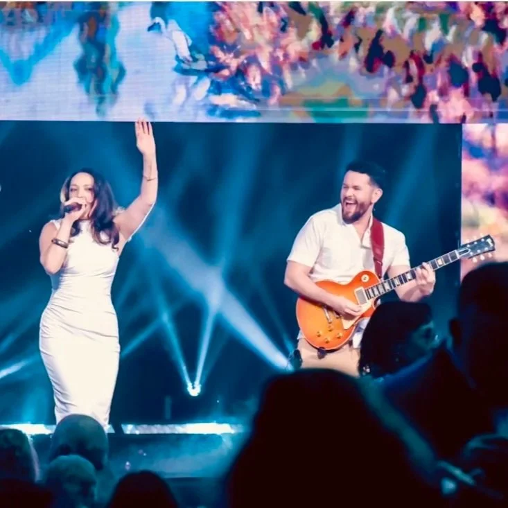 Female singer in a white dress performs beside a smiling guitarist on a brightly lit concert stage.