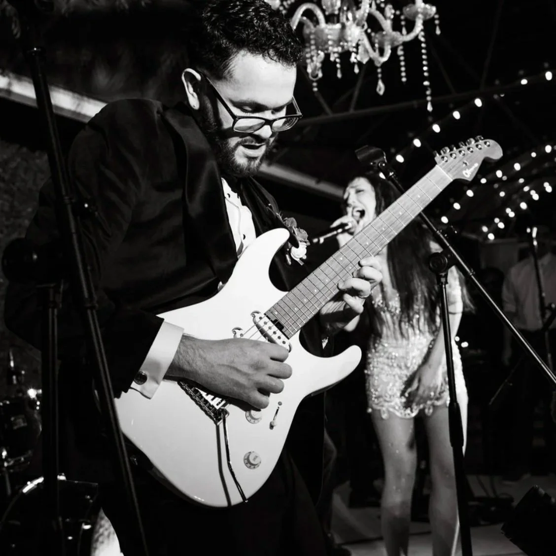 Black-and-white photo of a guitarist performing onstage while a singer performs behind him under festive lights.